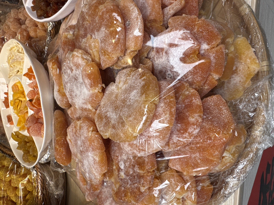 The scene depicts a market stall or shop display in Furong, China, presenting an array of candied and dried fruits. In the immediate foreground, a substantial pile of irregularly shaped, flattened orange-brown pieces of candied fruit, possibly ginger, is visible. These pieces are coated with a white, crystalline powdery substance, consistent with sugar, and largely covered with clear plastic wrap. This main pile rests on a light-brown woven bamboo tray.
In the mid-ground, a white multi-compartment serving dish holds various other candied fruits. One section contains yellow-green, elongated pieces. Another holds smaller, reddish-orange, sugar-dusted fruits. A third section displays slender, pale yellow strips, likely candied citrus peel. To the right of this dish, a smaller white bowl contains dark brown, shriveled items resembling raisins or dried berries. Further to the upper left, a darker woven basket contains a mound of rounded, translucent yellow candied fruits, also partially covered in clear plastic wrap. No individuals are present in the frame, and no specific actions or interactions are occurring. The environment suggests an indoor retail setting with bright, uniform illumination.