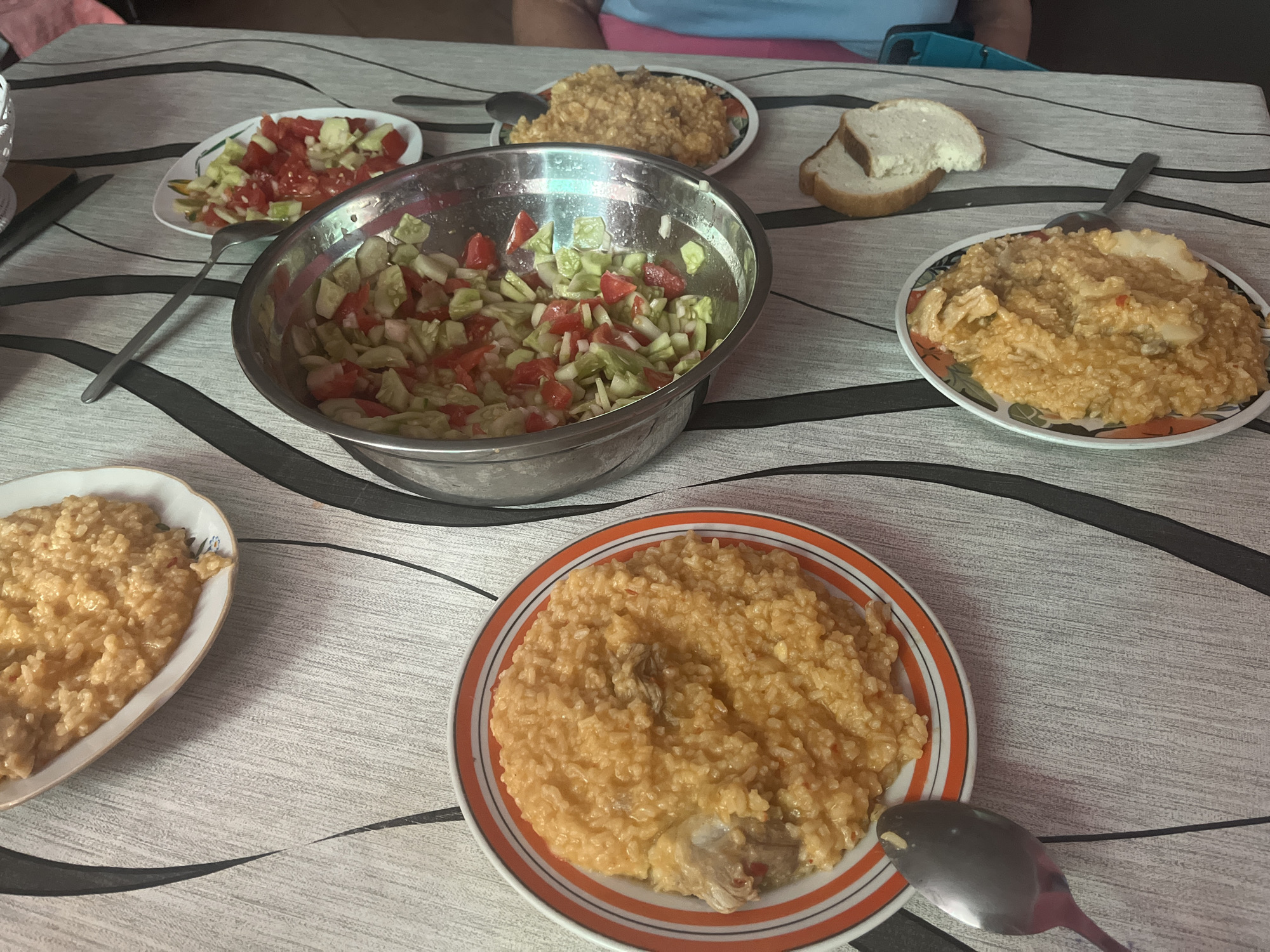 The image depicts a table set for a meal, likely in a home in Pivniceni, Moldova. The table is covered with a patterned tablecloth. Several dishes are visible, including plates of rice with meat, a large bowl of chopped salad containing tomatoes and cucumbers, and a plate of bread. Utensils, such as spoons and forks, are also present. In the background, part of a person wearing a blue shirt and pink shorts is visible. The scene appears to be a casual, home-cooked meal.