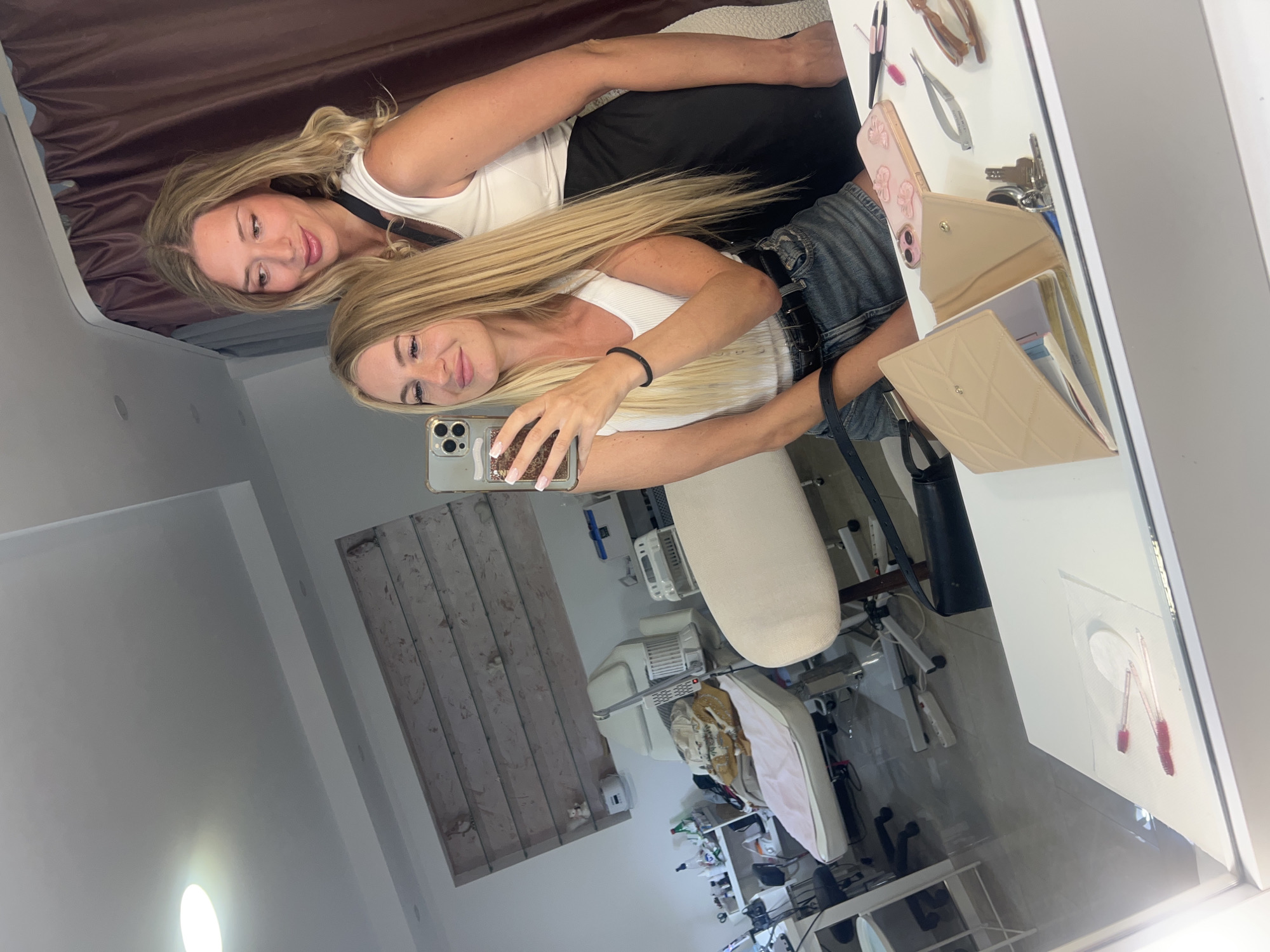 The image depicts two blonde women taking a selfie in a brightly lit, modern salon or spa in Budva, Montenegro. The woman in the foreground, wearing a white tank top and denim shorts, holds a smartphone and smiles at the camera. Behind her, a second woman, also with blonde hair, wears a white tank top and a black skirt. They are positioned in front of a large mirror that reflects a room containing salon equipment, including a massage table, various bottles and containers on shelves, and other beauty treatment tools. On the surface reflected in the mirror, which appears to be a counter, are a wallet, keys, tweezers, a phone case, and other small objects. The overall impression is a casual, candid moment captured within a professional setting.