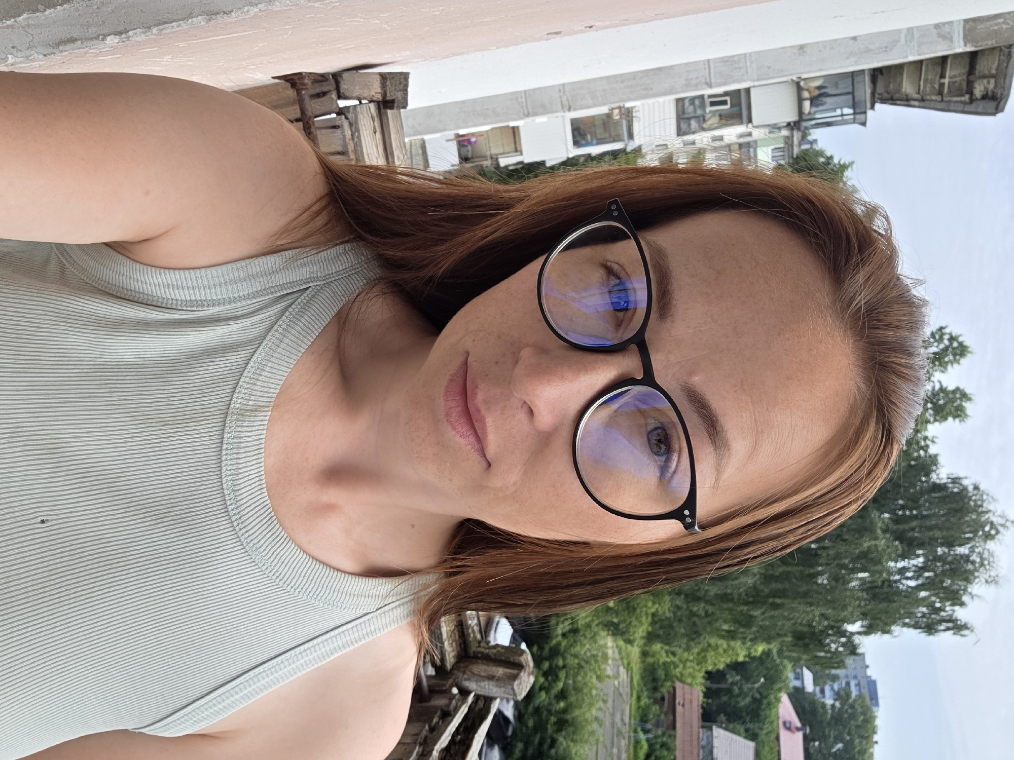 This is a close-up, self-portrait of a Caucasian woman with auburn hair and freckles. She is wearing black-rimmed glasses and a light green ribbed tank top. The woman is standing on a balcony, as indicated by the weathered wooden railing and concrete wall to her left. In the background, several multi-story buildings and trees are visible, suggesting an urban or suburban setting. The sky is overcast. The scene appears to be in the city of Chaikovsky, Russia, given the architectural style of the buildings and the foliage.