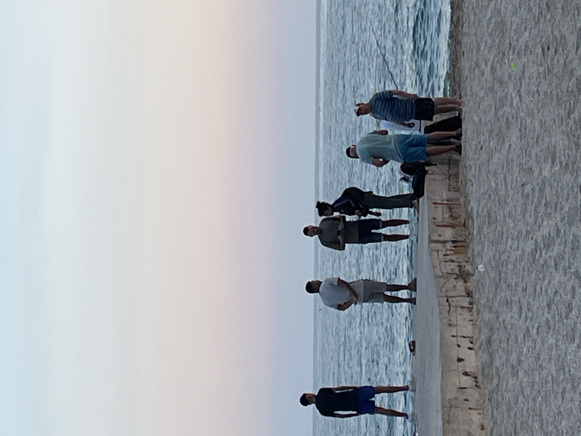 The image captures a serene coastal scene, likely in Alanya, Turkey, during either dawn or dusk. The sky, which occupies the upper left portion of the image, displays a soft gradient from light blue to a faint pinkish-orange, indicating the gentle light of early morning or late evening.
The main subjects are approximately seven to eight individuals, predominantly men, scattered along a concrete pier or embankment that extends into a calm body of water. Most of the individuals are casually dressed in t-shirts and shorts. Several people are engaged in recreational fishing, holding fishing rods that extend over the water, which shows subtle ripples. One individual in the middle appears to be a woman or a person with long hair, bending down or crouching near the edge of the pier.
To the right of the concrete structure, a sandy or gravelly area is visible, suggesting a beach or foreshore adjacent to the pier. A small, bright yellow ball is noticeable on this sandy ground. The concrete structure itself appears sturdy but shows some texture and wear. The overall atmosphere conveyed is one of peaceful leisure and outdoor activity. There is no visible text in the image.