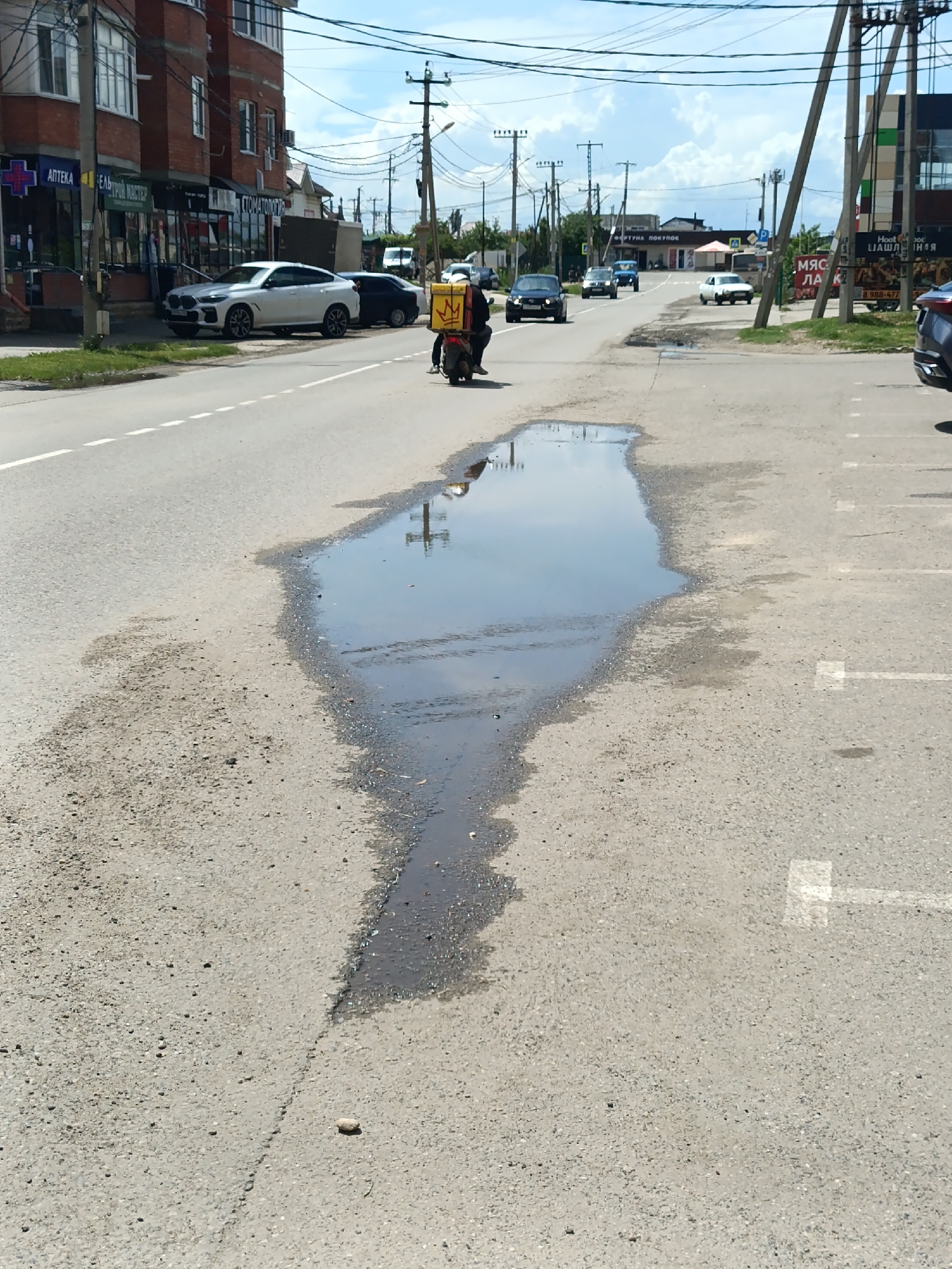 The image, taken in Yablonovskiy, Russia, depicts a street with a large puddle in the middle. The puddle reflects the sky and nearby objects, including power lines. A motorcyclist with a delivery box is traveling on the road ahead of the puddle. Several parked cars are visible on the side of the street, including a white SUV. Buildings line the street, housing businesses such as a pharmacy (АПТЕКА) and a dental clinic (СТОМАТОЛОГ). Parking spaces with painted markings are present on the right side of the road. Other vehicles are driving on the road in the distance. Power lines and poles run along the street.