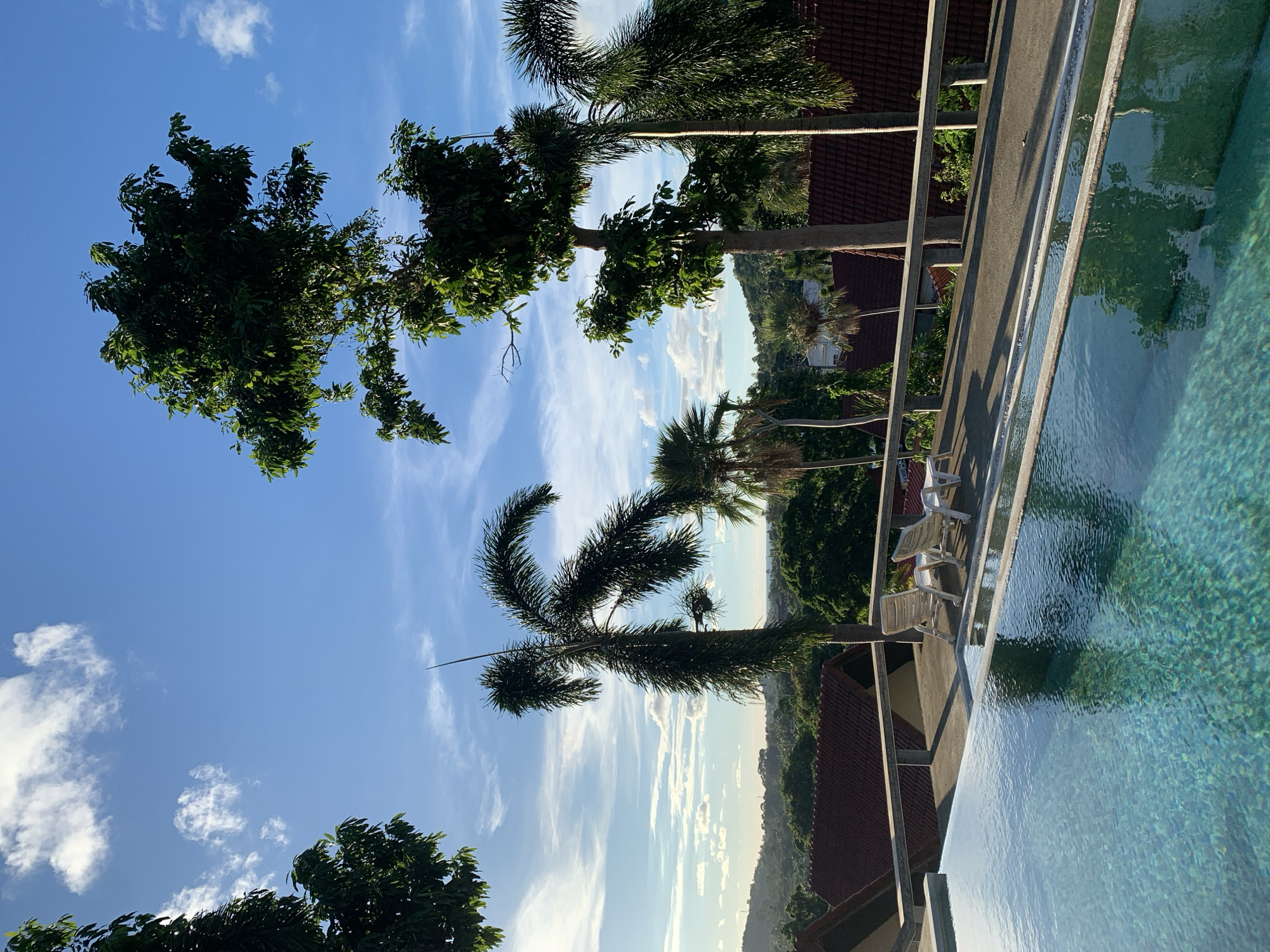 The image depicts an outdoor swimming pool with clear blue water, reflecting the sky and surrounding greenery. Two white lounge chairs are positioned on the pool deck, overlooking a tropical landscape. The background features lush vegetation, palm trees, and houses with red tiled roofs, suggesting a warm, possibly humid climate. The sky is a vibrant blue, dotted with wispy white clouds. The overall impression is one of a serene and luxurious resort setting, likely in or around the coastal area of Ban Bang Rak, Thailand, a region known for its beaches, resorts, and natural beauty.
