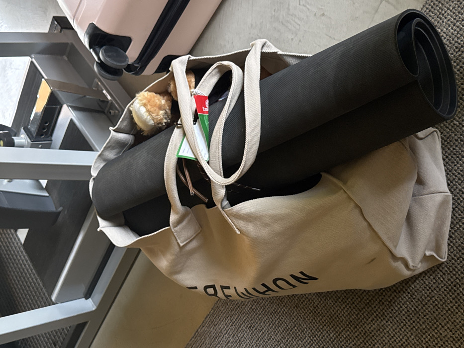 A beige tote bag lies on the floor, packed with a rolled-up black yoga mat, a stuffed animal, and a green tag attached to the strap. The bag is placed next to a gray, multi-tiered metal structure and a pink suitcase. The bag has "NDHMMON" printed on it, but the text is mirrored. The floor is covered in a carpet that appears to be gray.