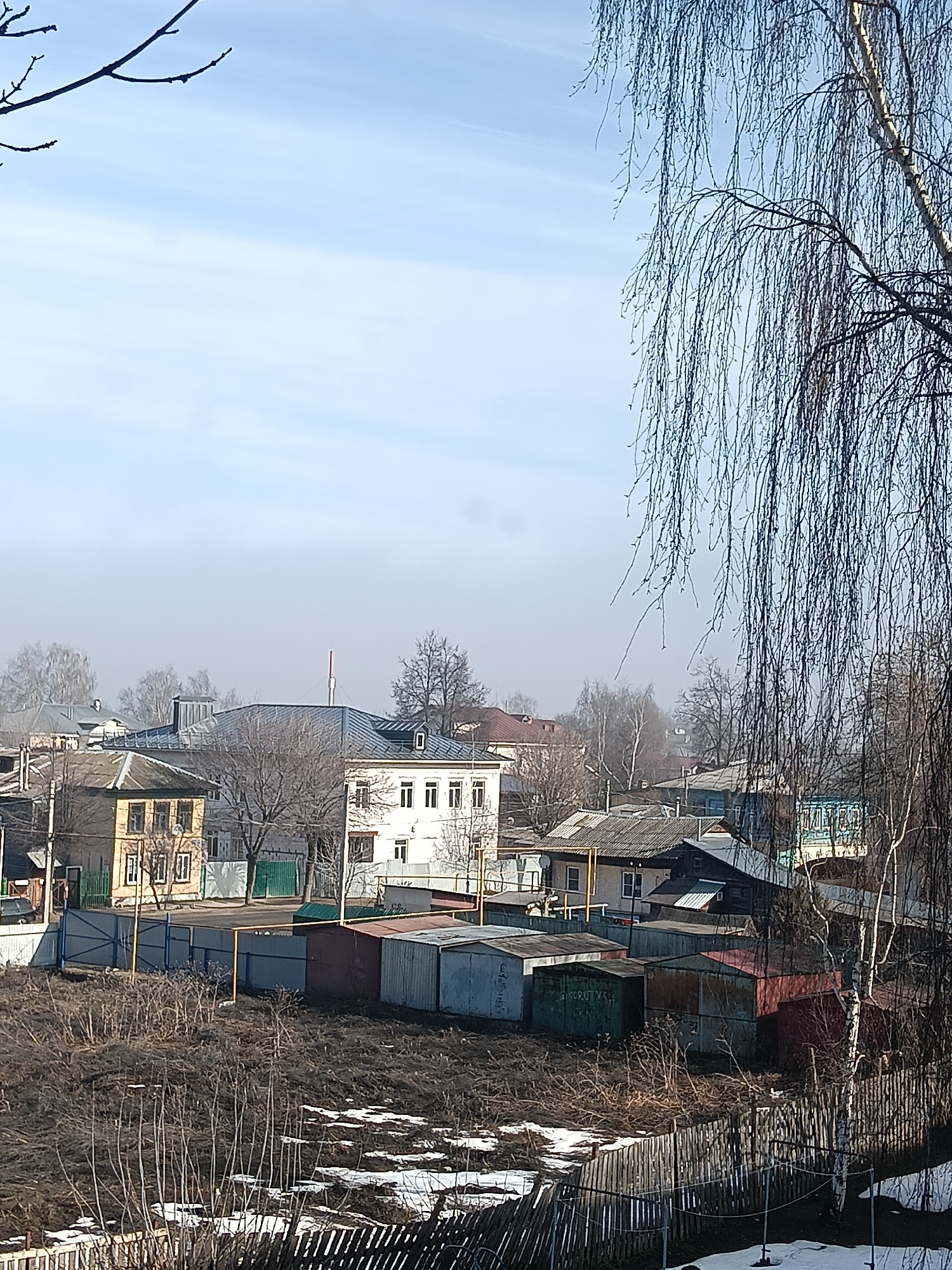 The image depicts a cityscape in Murom, Russia, likely taken in early spring. In the foreground, a dilapidated wooden fence partially obscures a patch of barren ground with scattered snow patches and dried vegetation. Beyond the fence, a row of metal garages of varying colors (gray, green, red) line the edge of a street. Further in the background, several buildings are visible, including a two-story yellow wooden structure and a three-story white building with a dark roof and a red chimney. Bare trees are scattered throughout the scene, with a prominent birch tree on the right side of the frame. A bare tree branch appears in the upper left corner. The sky is pale blue with a hazy overcast. No people are visible.