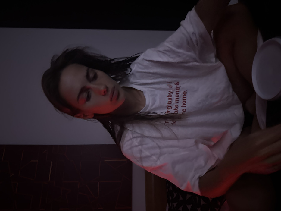 A young woman sits indoors. She has dark hair and is wearing a white t-shirt. Text is printed in red on the shirt. Her eyes are closed. The scene is lit with reddish light. A white plate-like object is visible near her right hand. A wall or headboard featuring a geometric pattern is behind her and to her left. The background colors are muted tones of gray, black, and white.
