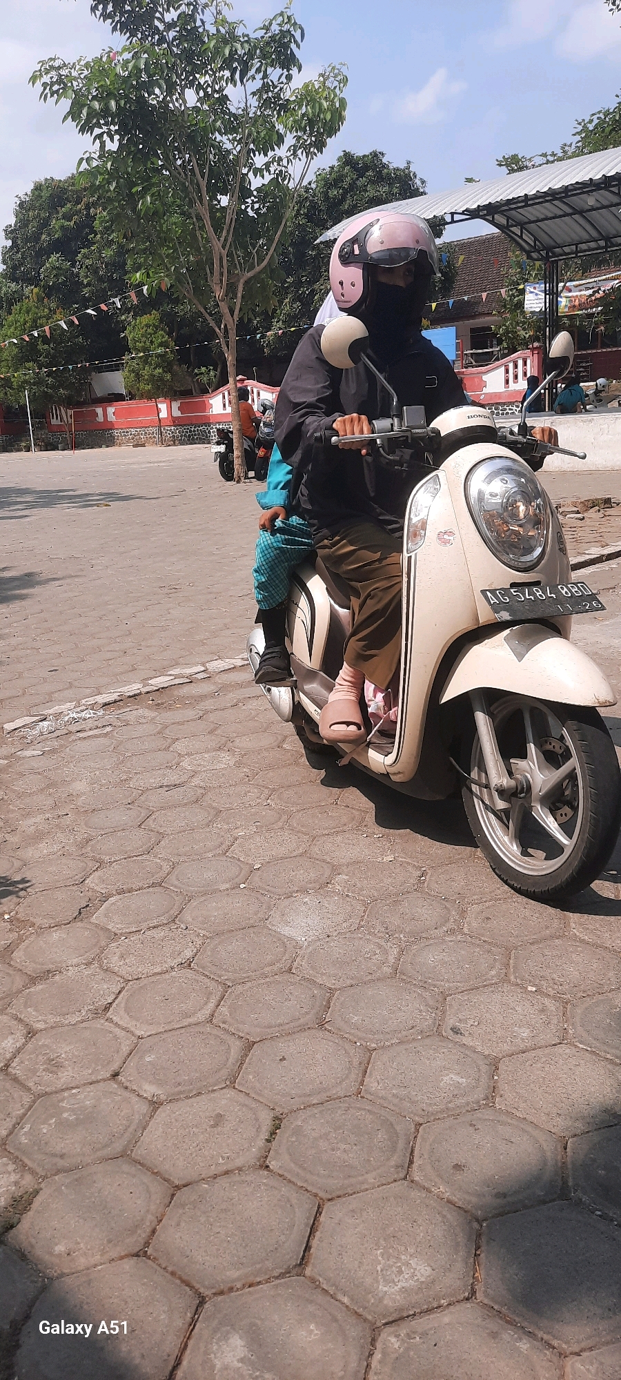 In the city of Tangkil, Indonesia, two individuals are seated on a light-colored scooter. The person in front, wearing a black jacket, a pink helmet, and a black face mask, is operating the scooter. The individual behind them is wearing a blue and green checkered garment. The scooter is parked on a hexagonal-patterned paved surface, with a street in the background and trees and foliage contributing to the outdoor setting. The license plate of the scooter reads "AG 5484 BBD".