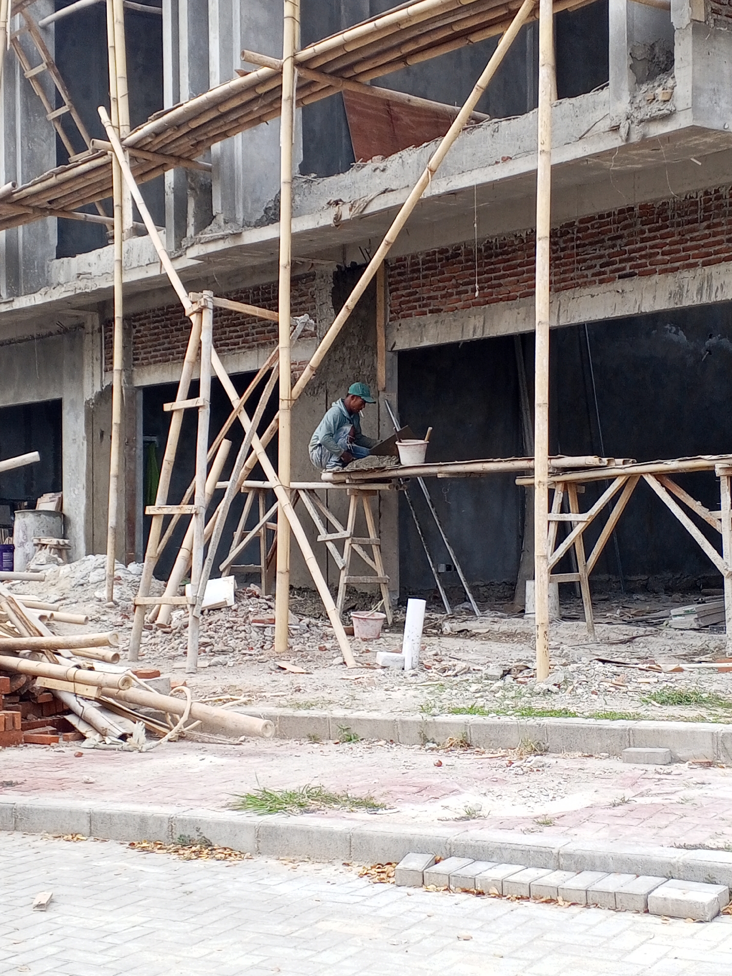 An outdoor, eye-level, vertical shot captures a construction scene in Serang, Indonesia. A person wearing a green cap and a light blue long-sleeved shirt is crouched on bamboo scaffolding, working with tools and a bucket on a concrete platform. Surrounding the individual and extending into the foreground and background is a complex structure of bamboo scaffolding, building materials like bricks and concrete debris, and partially constructed building walls. The ground consists of loose debris and some paving stones in the lower portion of the frame. The overall atmosphere is that of an active construction site.
