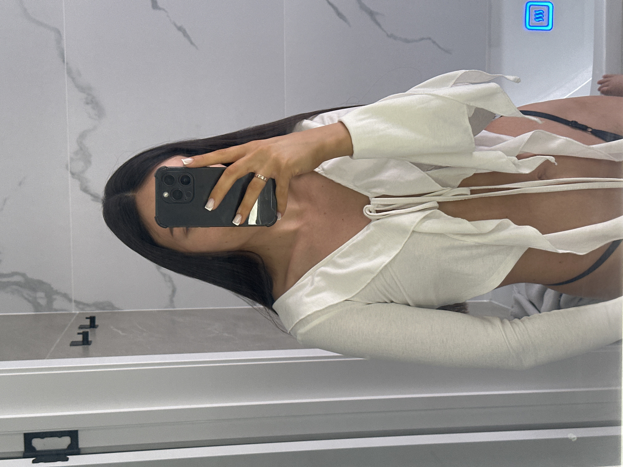A young woman is depicted taking a mirror selfie in a modern bathroom. She has long, dark, straight hair parted down the middle, with her face mostly obscured by the black-cased smartphone she holds in her left hand. Her right hand, featuring French-tipped nails and a narrow gold-toned ring on the ring finger, also grips the phone. She is wearing a cream-colored, long-sleeved crop top with a ruffled V-neckline and ties at the front, exposing her midriff. A strap of dark, thong-style underwear is visible on her right hip. The bathroom features light gray and white marble-patterned tiles on the walls. To the left, a white-framed shower partition with a black handle is visible. Two black, L-shaped hooks are mounted on a plain light gray wall panel to the right of the shower. A white bathtub is partially visible in the bottom right corner, featuring a blue illuminated control panel with three wavy lines. The woman's right foot is visible, seemingly resting inside or near the tub. The location is an indoor bathroom; however, there are no specific visual indicators within the image to determine a precise city or country, such as Эстачи, Russia.