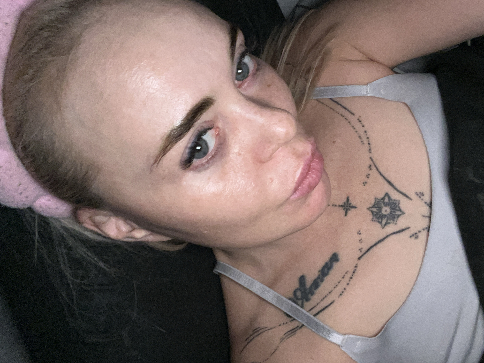 A woman with blonde hair is looking directly into the camera with her mouth slightly open. She is wearing a light gray bra and has tattoos on her chest. The background is dark and out of focus.
