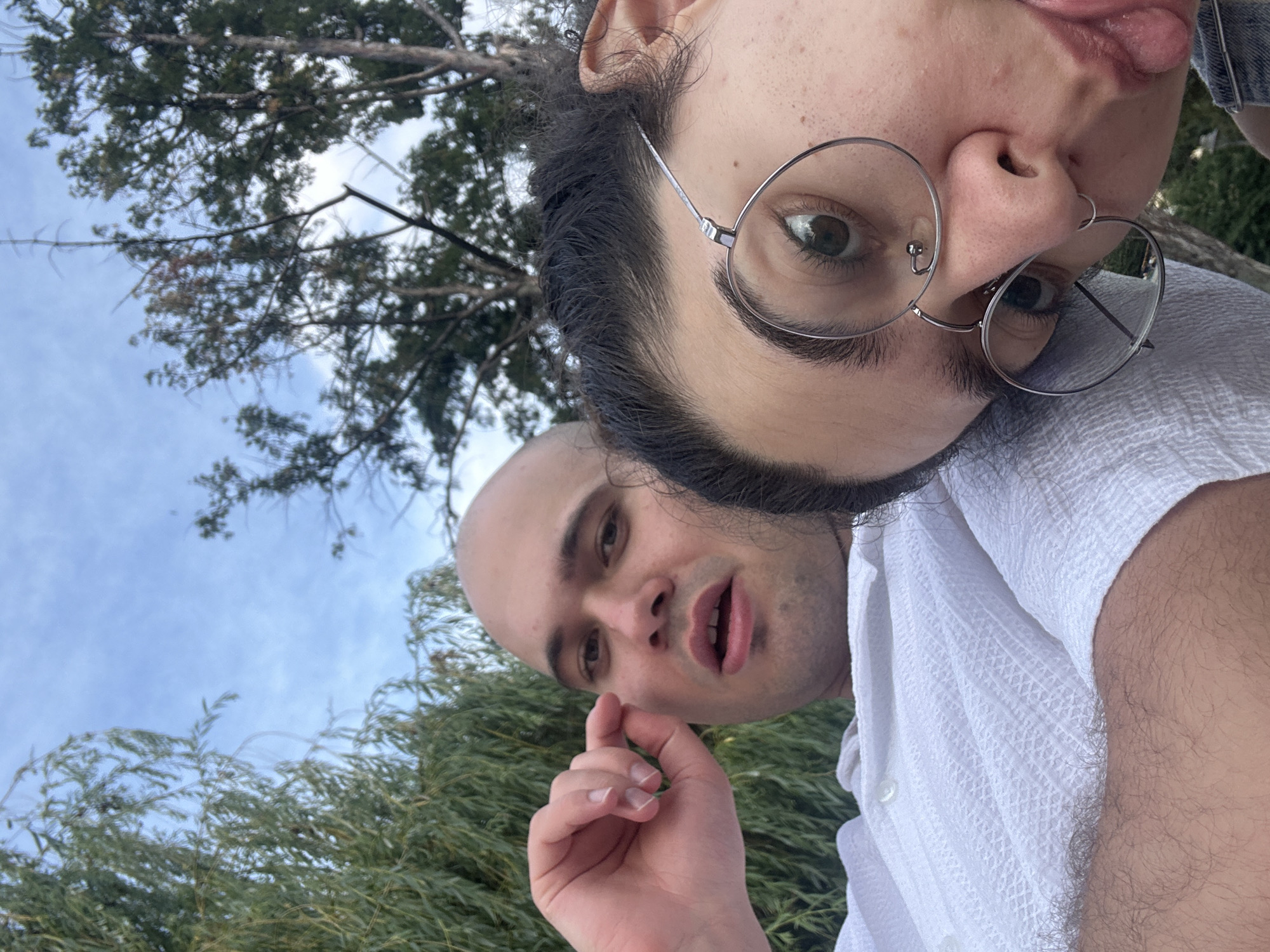 The media file is a selfie taken outdoors, with two individuals visible. One individual, positioned closer to the camera, has dark, curly hair and is wearing round-rimmed glasses. Their face is partially obscured, with only one eye and the nose visible. They appear to be sticking out their tongue. The other individual, positioned slightly further away, is bald and wearing a white, textured shirt. Their mouth is open, and their hand is raised near their mouth.
The background suggests an outdoor setting with greenery and a cloudy sky. The presence of trees and foliage indicates a natural environment, possibly a park or garden. The lighting suggests it is daytime, with diffused light, implying overcast or late afternoon conditions. The overall mood appears casual and playful, given the expressions of the individuals. No text is visible in the image. The location context is Sofia, Bulgaria, but this is not discernible from the image itself.