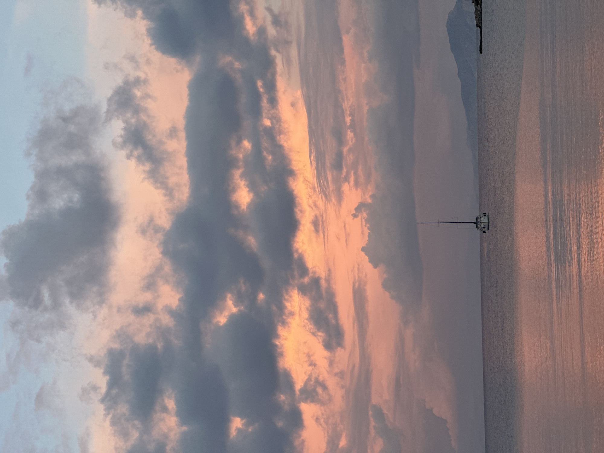 The image captures a vertical view of a seascape in Datça, Turkey, likely during sunset. A body of water fills the right half of the frame, reflecting pinkish hues from the sky. In the center, a white sailboat with a tall mast is anchored. In the background, a land mass or coastline can be seen. Dominating the left half of the frame is a dramatic sky filled with clouds colored in shades of gray, peach, and orange, suggesting either sunrise or sunset. There are no visible people in the image.