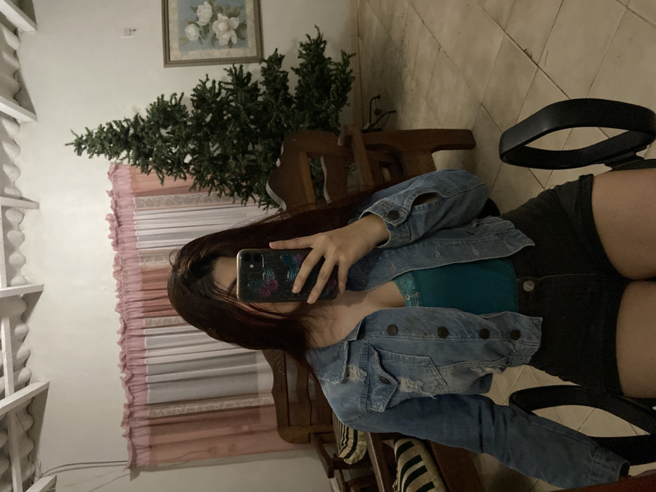 A woman in a denim jacket, green top, and black shorts sits in a chair, her face obscured by a cell phone as she takes a selfie. Long, dark hair falls across her shoulder. Behind her, a room contains a pine tree-like plant, a painting of white flowers, a wooden chair, and a sheer pink and white curtain. The floor is tiled. The room is dimly lit.