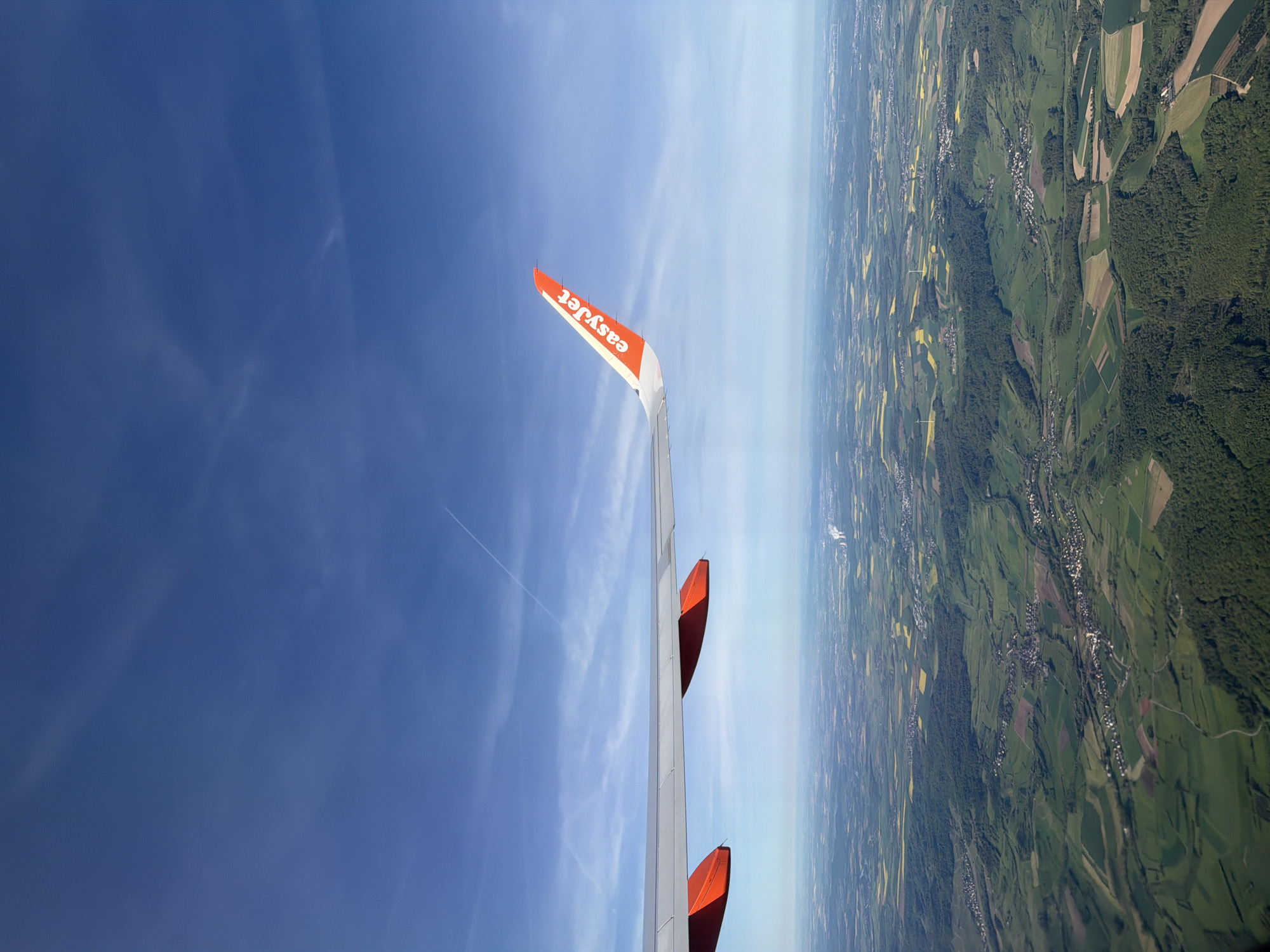 This image presents a vibrant aerial view captured from the window of an easyJet airplane, offering a clear perspective of the landscape below and the sky above during daylight hours. The scene is correctly oriented with the sky at the top and the ground at the bottom.
In the foreground, the clean white wing of the aircraft dominates the left side of the frame, extending towards the right. Its distinct orange winglet proudly displays the "easyJet" logo in white lettering, oriented such that it reads correctly from this viewpoint. Further along the wing, towards the center, two red/orange housing structures, likely for navigation lights or other wing components, are visible.
The upper portion of the image reveals a vast expanse of bright blue sky, largely clear but with subtle, wispy white clouds streaking across. A distinct, thin white contrail from another aircraft is visible in the upper-mid section of the sky.
Below the wing, the landscape unfolds as a rich tapestry of cultivated fields and dense woodlands. The fields vary in color, showing shades of vibrant green, brown, and patches of bright yellow, possibly indicating different crops like rapeseed or wheat. Numerous clusters of buildings suggest small towns or villages nestled among the agricultural land and forests. A hazy horizon line separates the earth from the sky in the distance, indicating clear weather conditions.
The overall setting is an expansive, well-maintained rural European landscape, consistent with an aerial flight over regions like Milan, Italy, as indicated by the context. The time of day appears to be sunny, likely morning or afternoon.