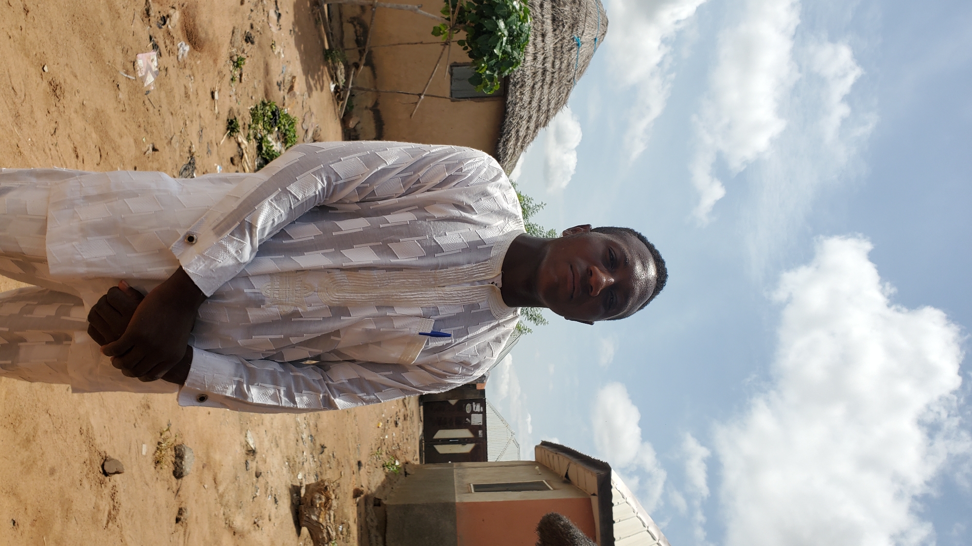A medium shot shows a young Black male standing outdoors in what appears to be a village in Jalingo, Nigeria. He is wearing a white, long-sleeved traditional garment with a geometric pattern and a blue pen tucked into the left breast pocket. His hands are clasped in front of him. Behind him, to the left, is a circular hut with a thatched roof and a small tree. To the right, a building with a concrete foundation and a corrugated metal roof is visible. The ground is sandy and uneven, with scattered debris. The sky is bright blue with scattered white clouds.