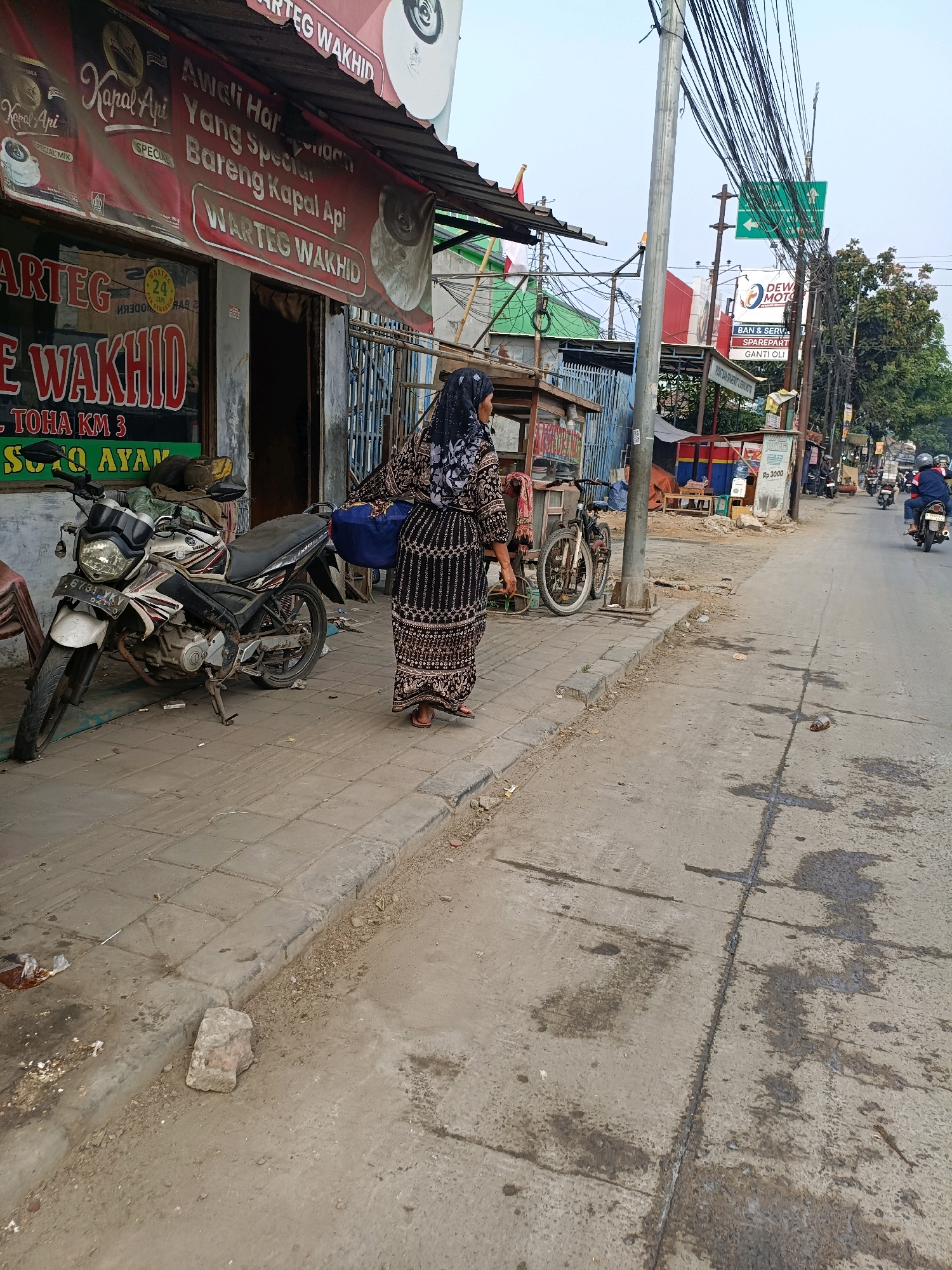 The image depicts a street scene in Tangerang, Indonesia, likely a commercial area with various shops and food stalls. A woman wearing a patterned hijab and a long, patterned dress stands on a sidewalk, facing away from the camera. She is carrying a blue bag. To her left, a motorcycle is parked on the sidewalk. A shop awning with "WARTEG WAKHID" written on it is visible behind the motorcycle. In the background, utility poles with numerous wires crisscross the sky, and a green traffic sign points to the left. Several other food stalls and shops line the street, with some customers and motorcyclists visible further down the road. The environment appears to be a typical urban street with paved sidewalks and a road surface showing signs of wear and water stains.
