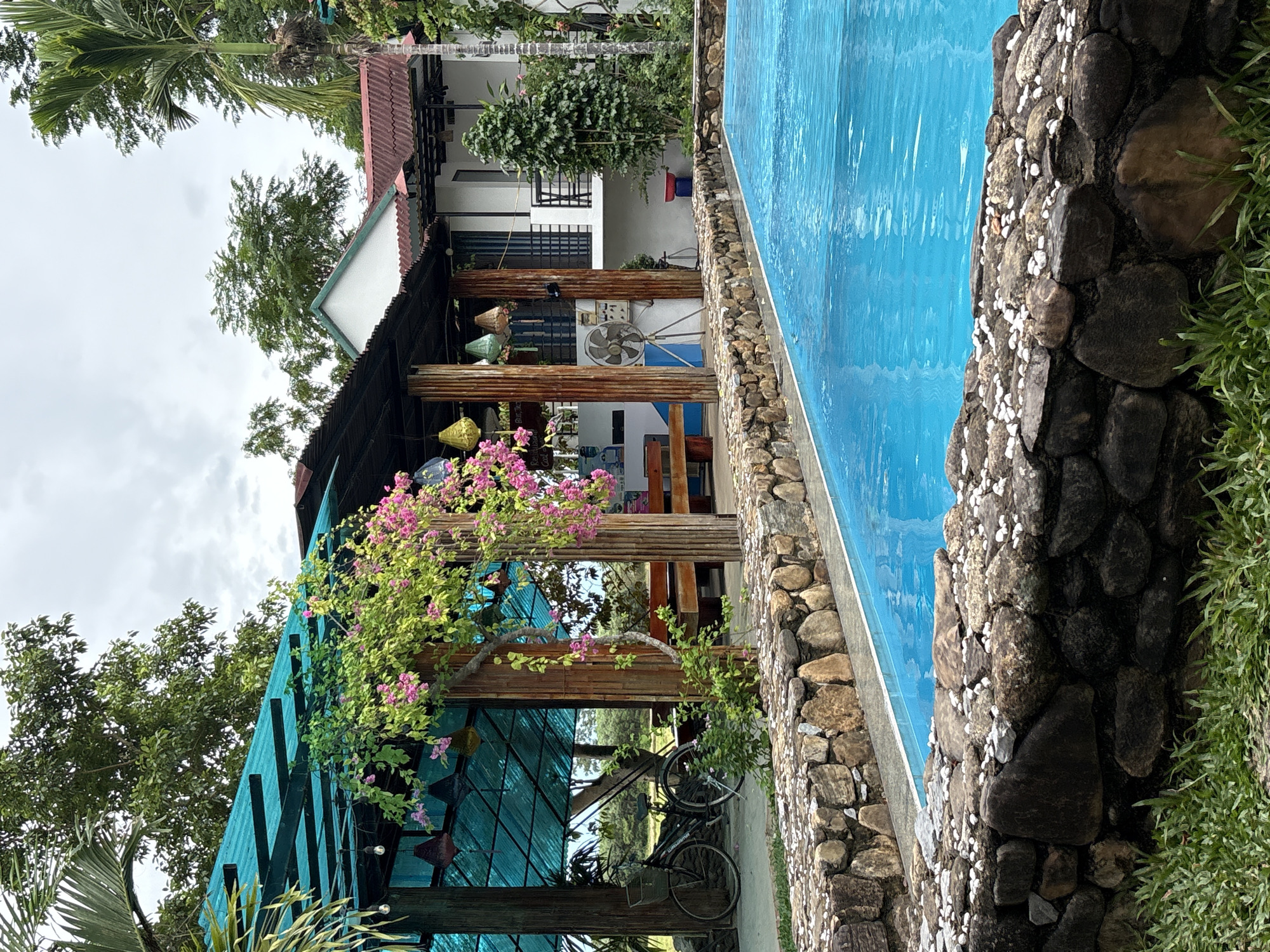 The image depicts an outdoor scene in Bố Trạch Commune, Vietnam, featuring a swimming pool with vibrant blue water. The pool is bordered by a stone wall, with a layer of smaller white stones on top. Beyond the pool, a covered outdoor seating area with a turquoise roof is visible, supported by wooden and corrugated metal posts. Pink bougainvillea vines are draped over the structure. In the background, a two-story building with a white facade and a reddish-brown roof is situated. Lush green trees and vegetation surround the area, with a clear sky overhead. A bicycle with a basket is parked to the left of the seating area. The overall impression is one of a tropical resort or guesthouse.