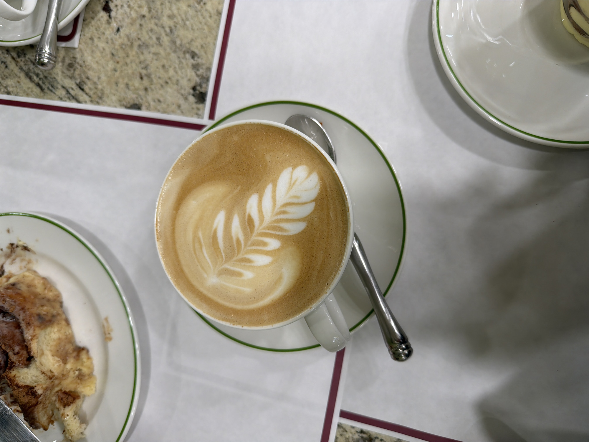 The image is a top-down view of a table setting in what appears to be a cafe or restaurant. Dominating the scene is a cup of latte with a feather-shaped latte art, placed on a saucer with a green rim. A spoon rests on the saucer next to the cup. To the left of the latte is a plate containing pastry. Another plate is partially visible in the upper right of the frame. Portions of a table runner or placemat are visible, with a red and white border. The table surface is white, except for sections in the upper left and lower right, which show a light, speckled texture, potentially granite. The setting suggests a casual meal. The city and country cannot be determined from the image alone.