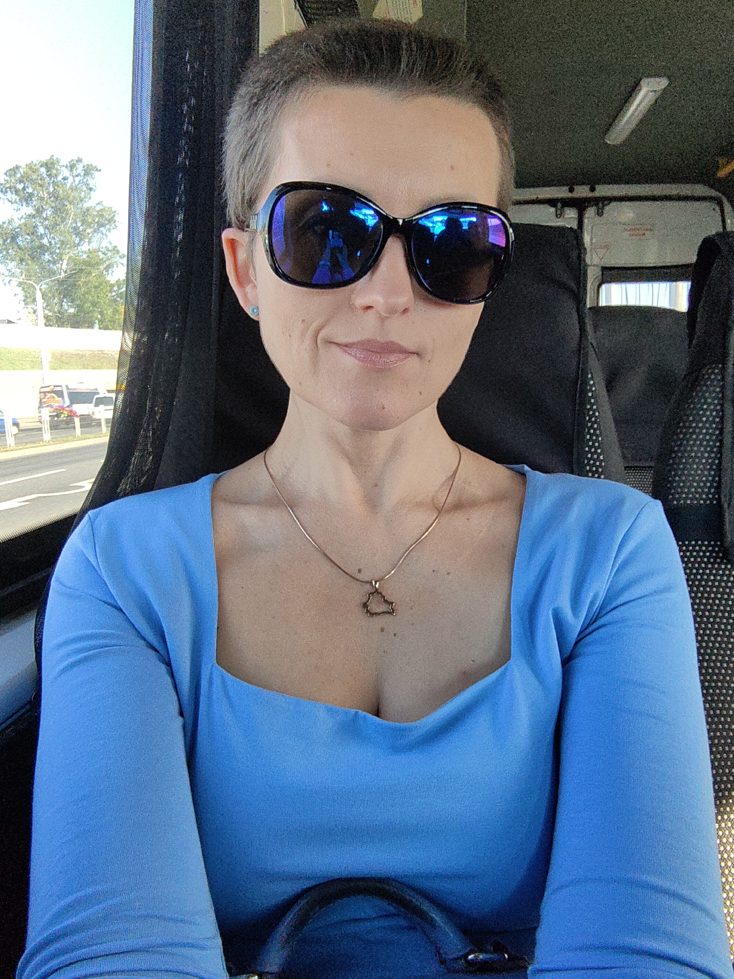 A woman with short, dark hair and light skin is seated in a vehicle, possibly a bus, as indicated by the seats and interior lighting. She is wearing large, dark-rimmed sunglasses with blue reflective lenses, a bright blue long-sleeved shirt with a square neckline, and a delicate gold necklace with a small, ornate pendant. She is also wearing small, light blue stud earrings. The background suggests an outdoor environment, with trees and a road visible through a window or opening to her left. The interior of the vehicle appears to be dark, with black seating. The lighting suggests it is daytime. The image is a selfie, taken from the chest up, with the woman looking towards the camera. There are no specific visual cues in the image that would place it in Minsk, Belarus.