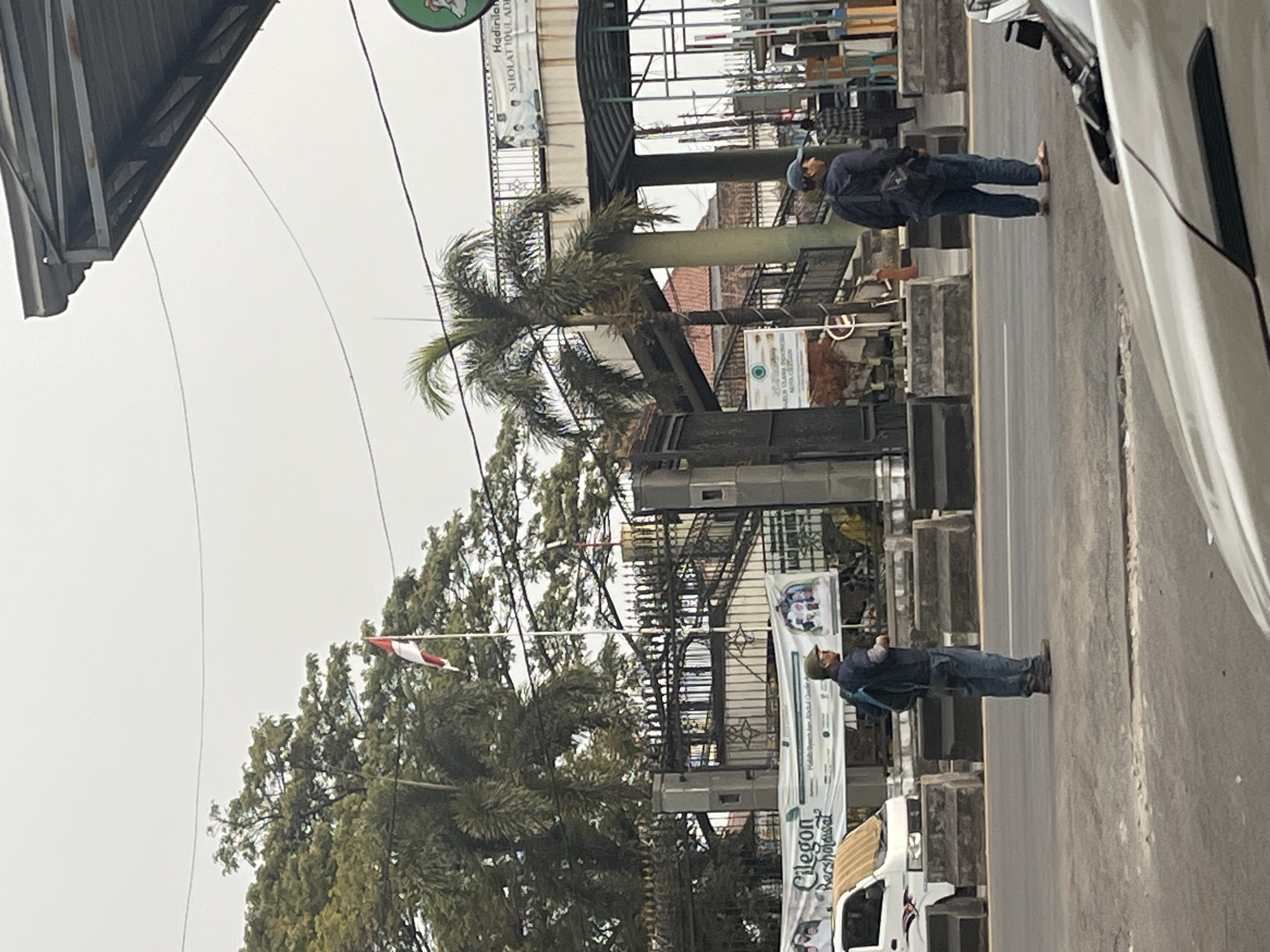 The image captures a street scene in Cilegon, Indonesia, on an overcast day. In the foreground, the hood of a light-colored car is visible. The mid-ground features a road with two men standing. The man on the left wears a dark blue shirt, blue jeans, and a green cap. He carries a dark backpack. The man on the right wears a dark blue jacket, blue jeans, and a blue cap. He also carries a dark backpack. Between the two men, a white van is parked. In the background, there is a large gateway with a fence, and a banner displaying text, including "Cilegon Bersholawat." A flag, red and white, flies on a pole above the gateway. Beyond the gateway, there is a building with a prominent staircase and an elevated walkway. Palm trees and other foliage are visible, interspersed with structures. The overall environment suggests an urban setting with some greenery.