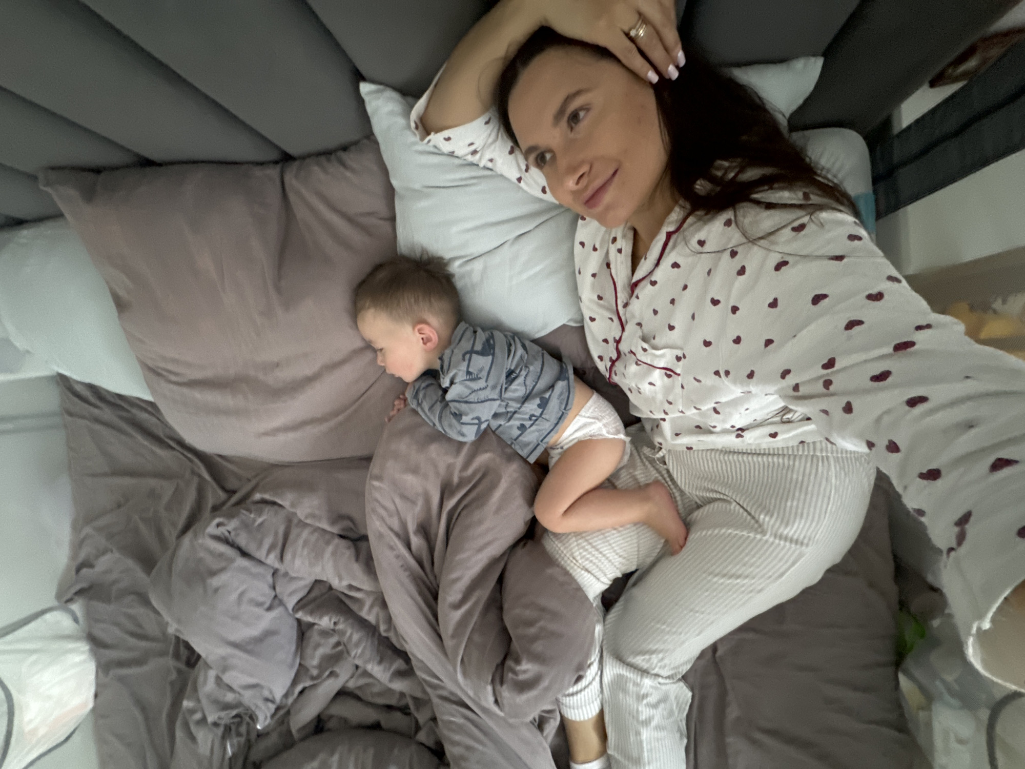 A woman and a child are lying on a bed. The woman is wearing a white pajama top with red hearts and white striped pajama bottoms. She is lying on her left side, with her right arm raised above her head. She is looking to her left. The child is lying on their stomach next to the woman, with their head resting on a pillow. The child is wearing a gray shirt with a dinosaur pattern and a diaper. The bed has a gray headboard and is covered with a gray sheet and pillows. The location is likely a private residence, but not specifically identifiable as being within the city of Odintsovo, Russia.