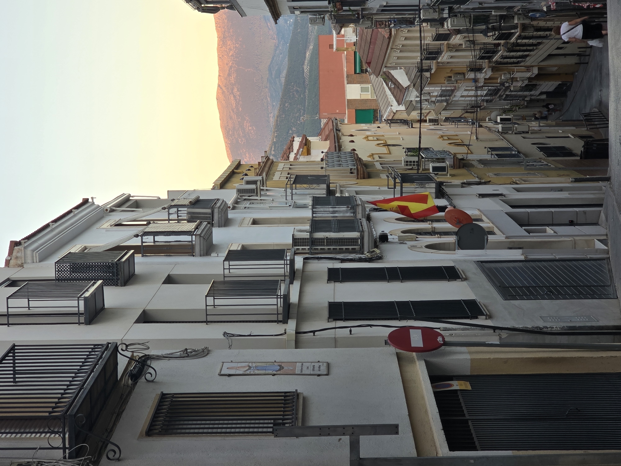 The image depicts a narrow, descending street in Jaén, Spain, characterized by tightly packed, multi-story buildings. The architecture features traditional balconies with black wrought-iron railings and numerous air conditioning units mounted on the facades. On the left side of the street, a white building displays a religious tile mosaic and a red "no entry" sign. A Spanish flag hangs prominently from a balcony on a yellow building further down the street. The street slopes downwards, revealing a vista of more buildings and a distant, sunlit mountain range. The sky is a gradient of pale blue and soft yellow, suggesting either dawn or dusk. Two women are visible in the distance at the bottom of the street, one walking away from the camera and carrying a white bag. The overall impression is of a steep, historic urban landscape bathed in warm, low light.