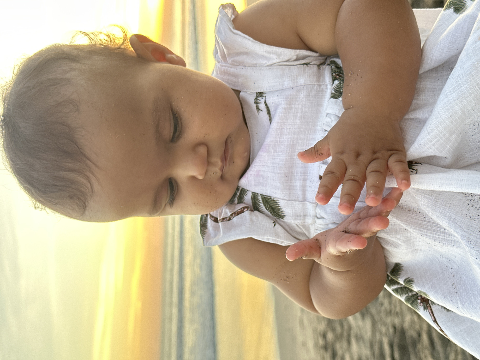 A baby is pictured in a white dress with a palm tree pattern, holding sand in their hands. The baby has brown hair and skin, and appears to be on a beach. The background suggests it is sunset. This photo was taken at North Kuta, Indonesia.