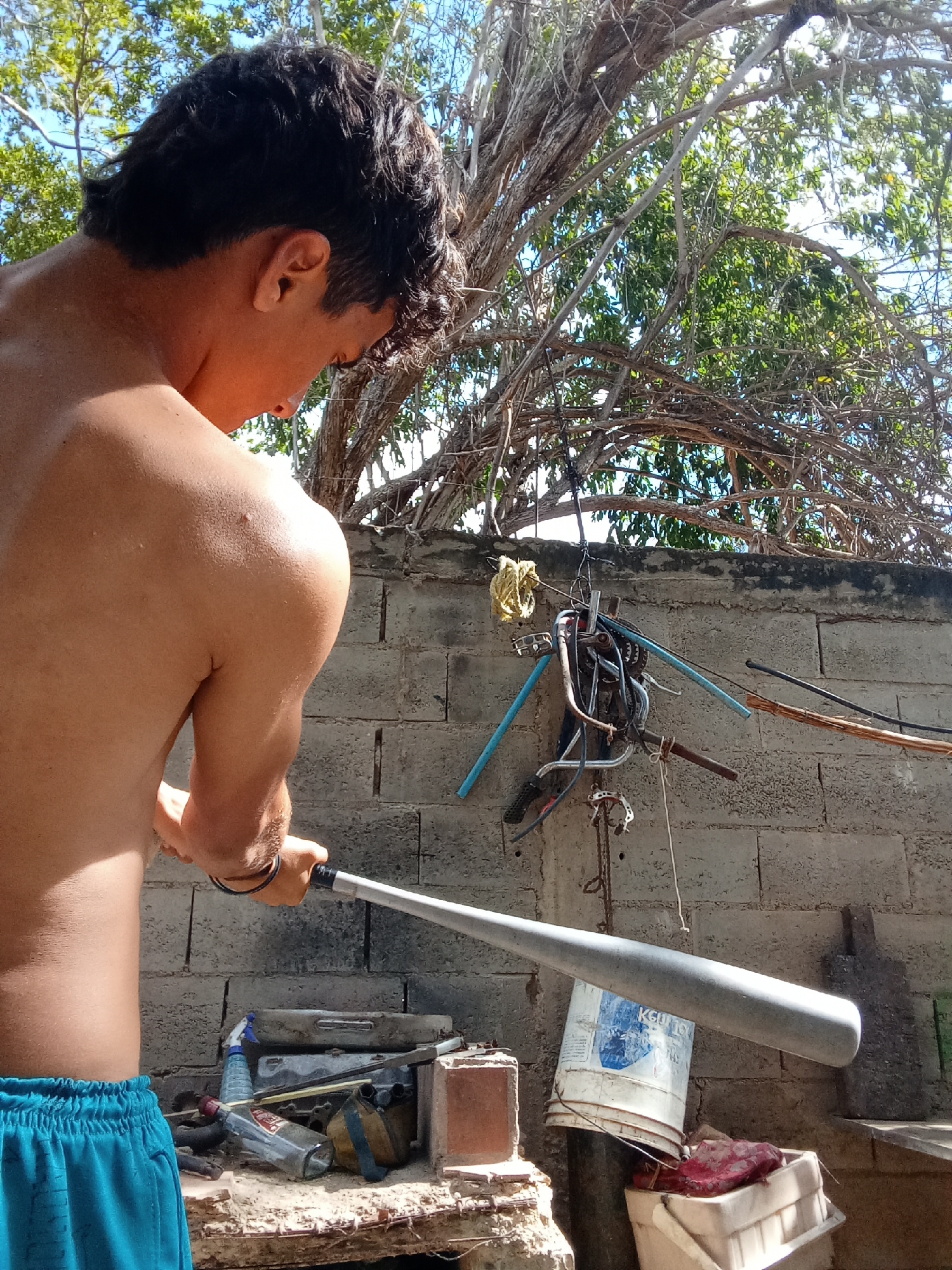 A shirtless man in Juan Griego, Venezuela stands in front of a cinder block wall, holding a gray metal baseball bat. The man is wearing blue shorts and faces away from the viewer, looking down at something out of view. Behind the wall are branches of a tree. Hanging on the wall are various objects, including parts of a bicycle. A weathered shelf below the wall contains an assortment of items, including bottles, cans, and a bucket.