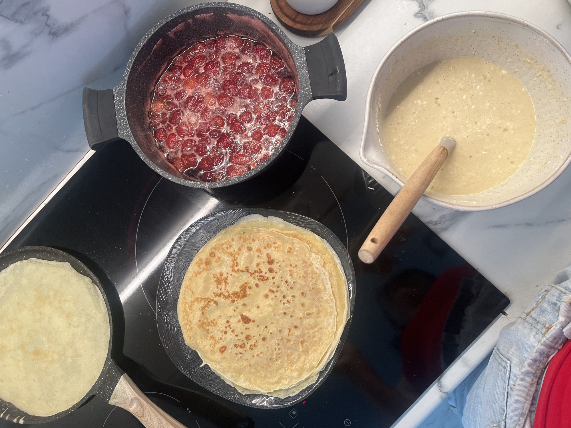 This overhead shot captures a scene of preparing crepes. In a dark grey pot on a black stovetop, a batch of strawberries is simmering, creating a vibrant red mixture. To the right, a large white bowl holds a pale yellow batter, with a wooden-handled whisk resting inside. On the stovetop, a crepe is being cooked in a dark pan on the left, and a stack of golden-brown crepes sits on a clear glass plate on the right. A wooden spatula with a hole in its handle is also visible, suggesting active cooking is in progress. The setting appears to be a modern kitchen with a white marble countertop. The lighting suggests it's daytime. The overall impression is one of domestic food preparation, with a focus on the ingredients and the process of making crepes. There is no visible text in the image. The location context provided (Aqsu city administration, Kazakhstan) is not visually evident from the image itself.