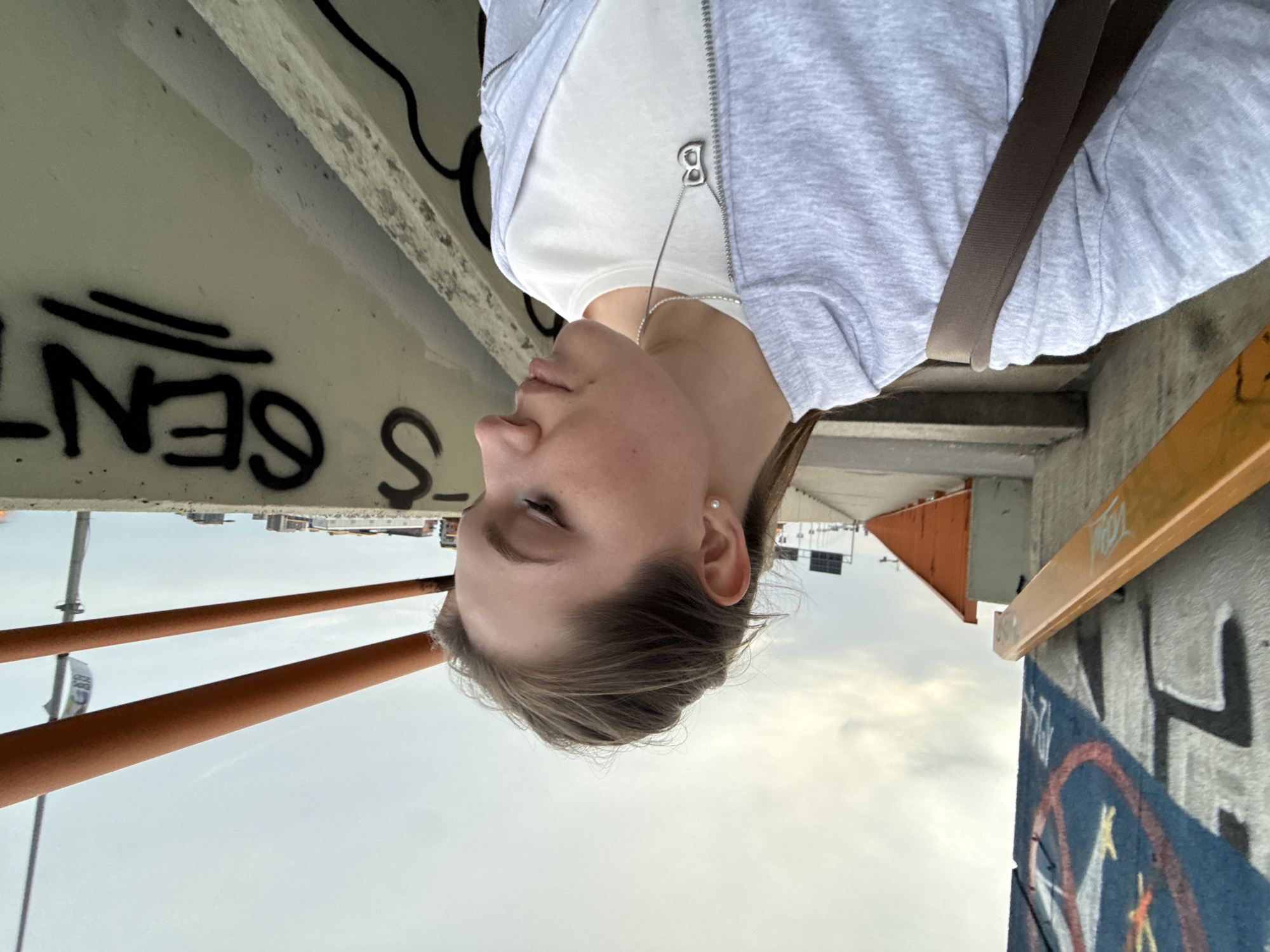A woman stands on a bridge in Belgrade, Serbia. She wears a white shirt under a grey zip-up jacket, with a brown shoulder strap across her body. Graffiti is visible on the concrete structures of the bridge. In the background, buildings are visible under a cloudy sky. The bridge railing is orange.