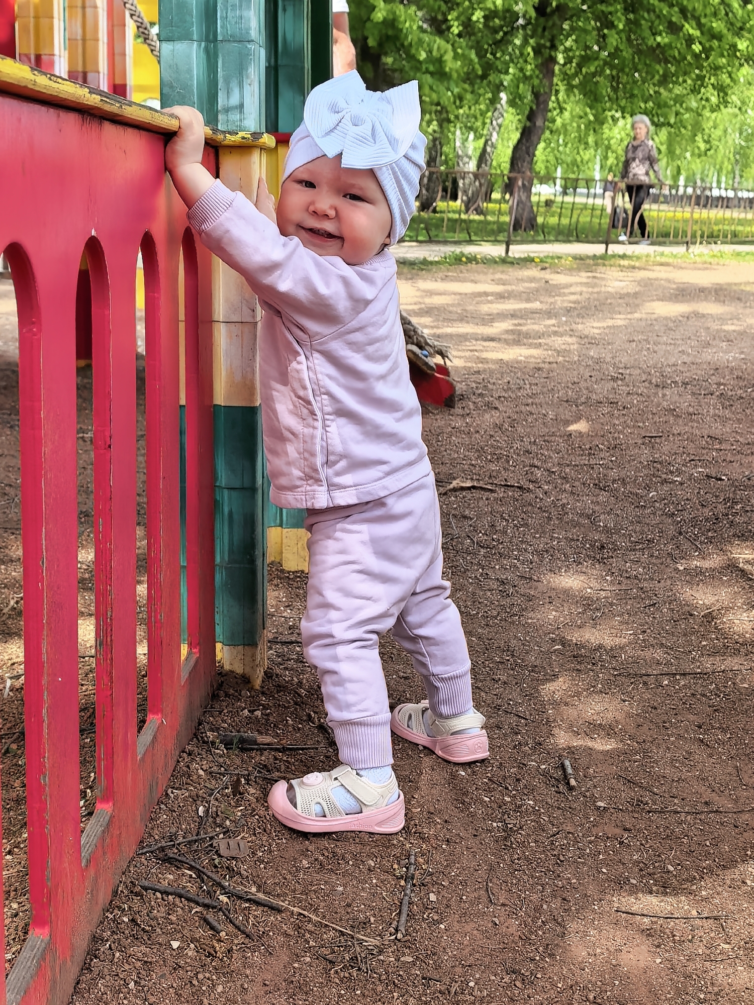 A young child stands on a mulch-covered ground in a park on a sunny day. The child, wearing a light lavender sweatsuit and a matching bow headband, is using both hands to hold onto a colorful, painted wooden structure, likely part of a playground. The structure has red vertical slats and a green and yellow vertical support post. The child is wearing pink and white sandals and smiling at the camera.
In the background, beyond the child and a small area of shade, there is a woman in a black and brown patterned top and black pants walking away from the camera on a paved path, partially obscured by trees and a metal fence. The background suggests a park-like setting with trees and vegetation.
The location is Sterlitamak, Russia, based on the image's context.