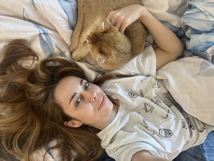 The scene is an indoor residential setting, located in the city of Bar, Montenegro. A person with light skin, long wavy reddish-brown hair, and light-colored eyes is depicted lying on their back in bed, looking towards the camera. The individual wears a white short-sleeved t-shirt featuring multiple black line-art graphics, including a beer can labeled "ŽIVELI," a dumbbell, a coffee cup, a cigarette, and text "SALUTE" and "KAKO." Their lower body is covered by white bedding. Positioned against the person's left side is a golden-brown domestic cat, lying on its side with its head oriented away from the camera. The person's left hand is resting on the cat's back. The bed is furnished with a duvet cover displaying an abstract pattern of blue and white wavy lines.