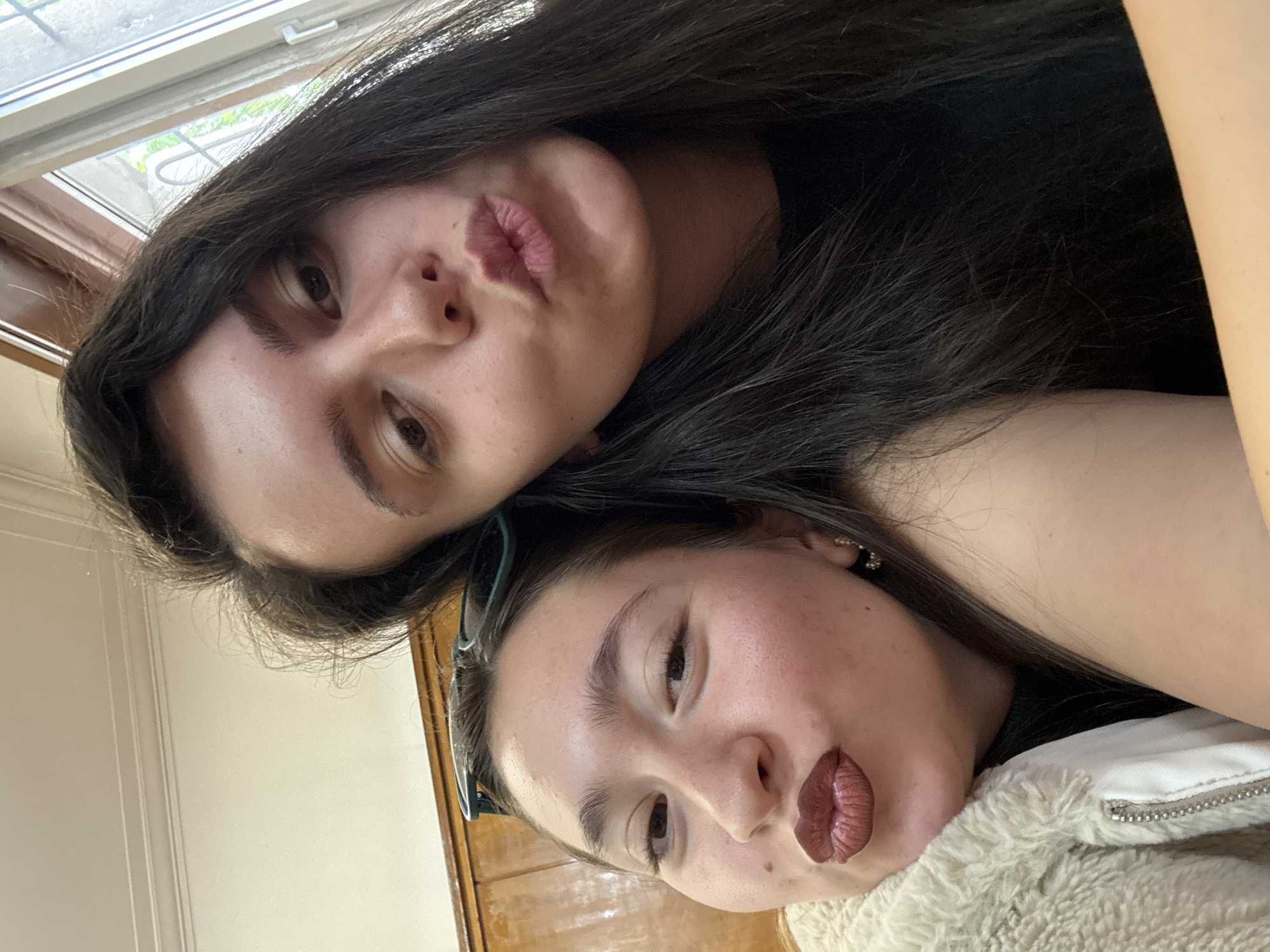 Two young women are posing closely together for a selfie. The woman on top has long, dark hair, light skin, and is making a kissy face. The woman underneath has her hair pulled back, is also making a kissy face, and is wearing a light-colored jacket. The background shows a window, a wooden door frame, and beige-colored walls. There is no evidence to suggest that the location is in Sofia, Bulgaria.