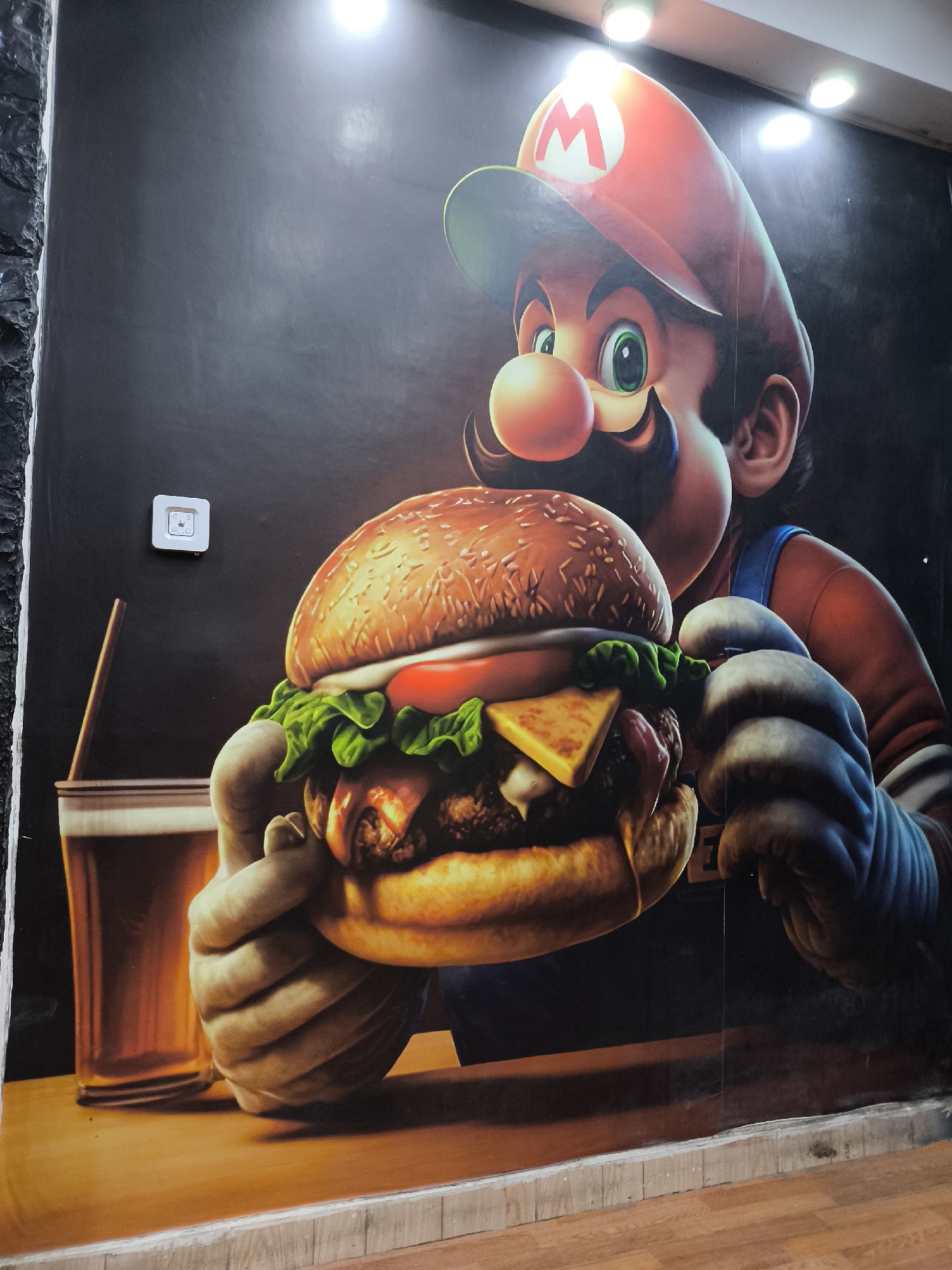 A mural on a wall depicts the video game character Mario holding a large hamburger. Next to the burger is a glass of liquid with a straw. A light switch is visible on the wall to the left of the burger. The wall is painted black. The image appears to be taken indoors, near the floor.
Location: Unknown, Unknown.