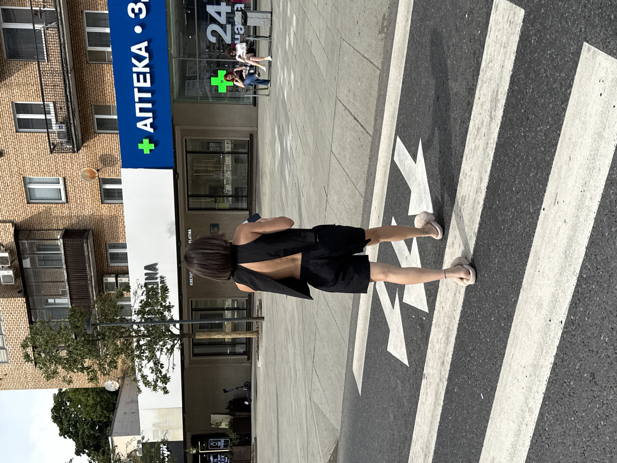 In Moscow, Russia, a woman with short dark hair, wearing a black sleeveless top with an open back and black shorts, walks across a pedestrian crosswalk. She is wearing fuzzy slip-on sandals. Behind her, a brick building with multiple windows and balconies is visible. A white awning stretches across the lower facade of the building. To the right, a bright blue sign displays "АПТЕКА" (Pharmacy) with a green plus sign. Further to the right, a glass storefront has a "24" displayed, indicating it is open 24 hours. Two young women are seated on a bench outside the pharmacy, one with red hair and the other with dark curly hair, both looking at their phones. The ground is paved with light-colored square tiles. The sky is overcast.