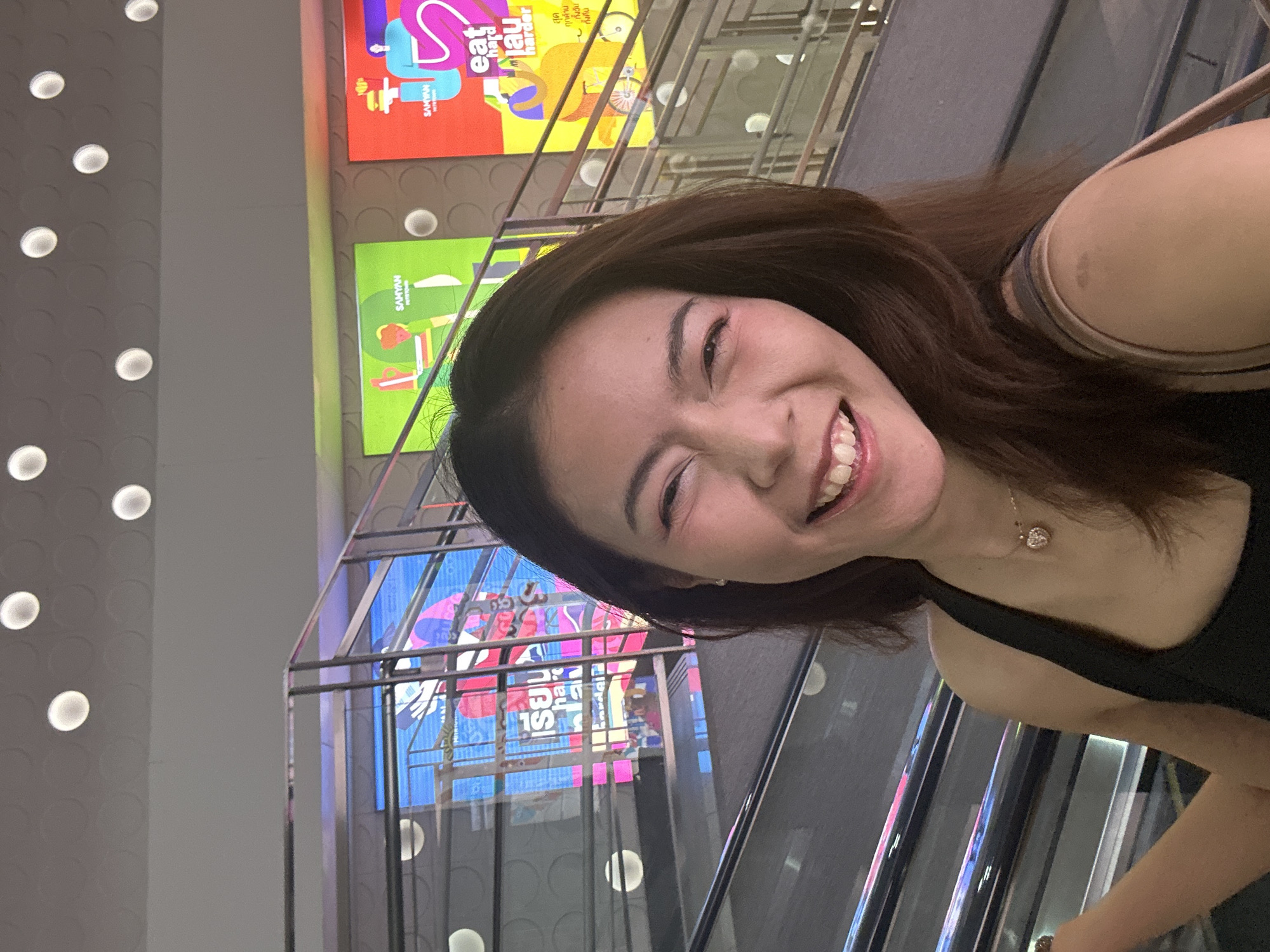 A young Asian woman with long brown hair is taking a selfie on an escalator in Bangkok, Thailand. She is wearing a black tank top and a heart-shaped necklace. She is smiling widely, with her eyes closed and her head tilted to the side. In the background, there are colorful digital advertisements displayed on a staircase. The overall scene is vibrant and modern, suggesting a lively urban environment.