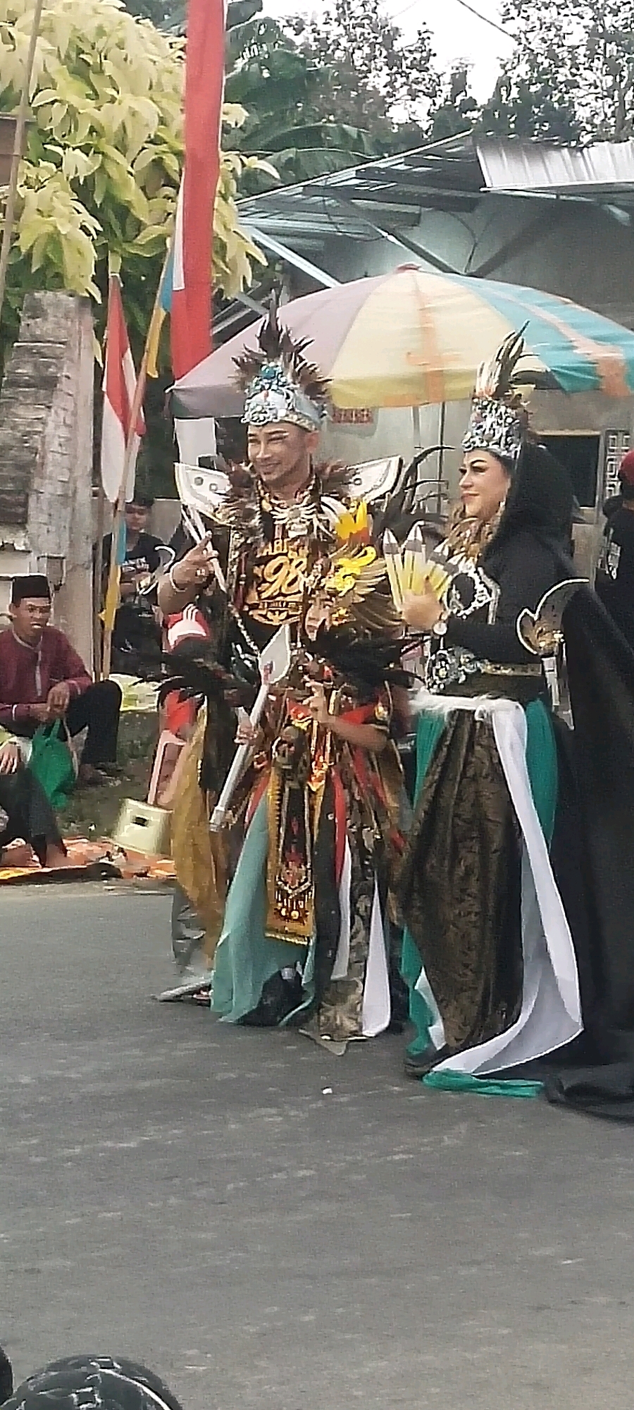 A medium shot captures a cultural performance in the city of Sabrang, Indonesia, with two individuals in elaborate costumes standing on a paved surface. The man on the left wears a headdress adorned with feathers and turquoise, a dark chest plate with gold accents and the number "98" prominently displayed, and a long, layered skirt in teal and gold. He holds a prop resembling a spear and is looking slightly to his right, with his left hand extended in a pointing gesture. To his right, a woman wears a similar headdress and a black hooded garment over a dress with a patterned brown skirt and a teal and white sash. She holds a fan of yellow, glowing objects in her left hand and is looking to her left. Behind them, a large umbrella with alternating yellow and green panels provides shade. To the far left, a man in a maroon shirt and black pants sits on the ground with a green bag beside him. In the background, a brick wall, lush green foliage, and trees are visible, along with a corrugated metal roof structure. The ground is a neutral gray asphalt surface, with several small white objects scattered on it. The overall atmosphere suggests a festive or ceremonial event.