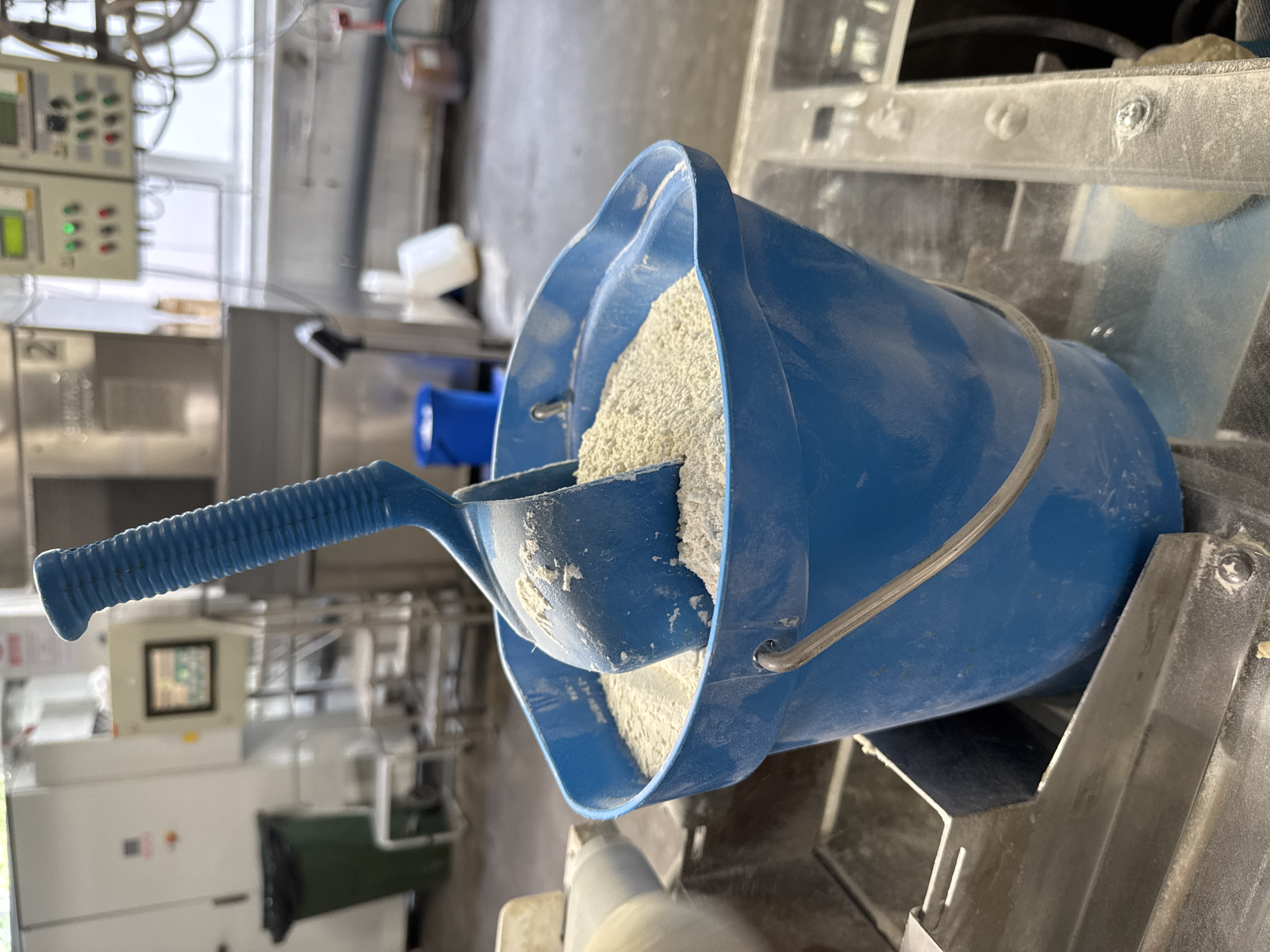 A close-up, slightly upward angle shot shows a blue plastic bucket filled with a light-colored granular substance, likely flour or a similar ingredient, in what appears to be an industrial setting. A blue plastic scoop with a ribbed handle is partially submerged in the substance, suggesting it was used for measurement or transfer. The bucket has a silver wire handle. The background is blurred but reveals various pieces of industrial equipment, including metal shelving, containers, and control panels with lights and screens. There is a green trash can to the left. The overall environment appears to be a food production facility or bakery, possibly in Dedovsk, Russia, given the mention of the location, though this cannot be confirmed from visual cues alone. The lighting is bright, indicating overhead industrial lighting.