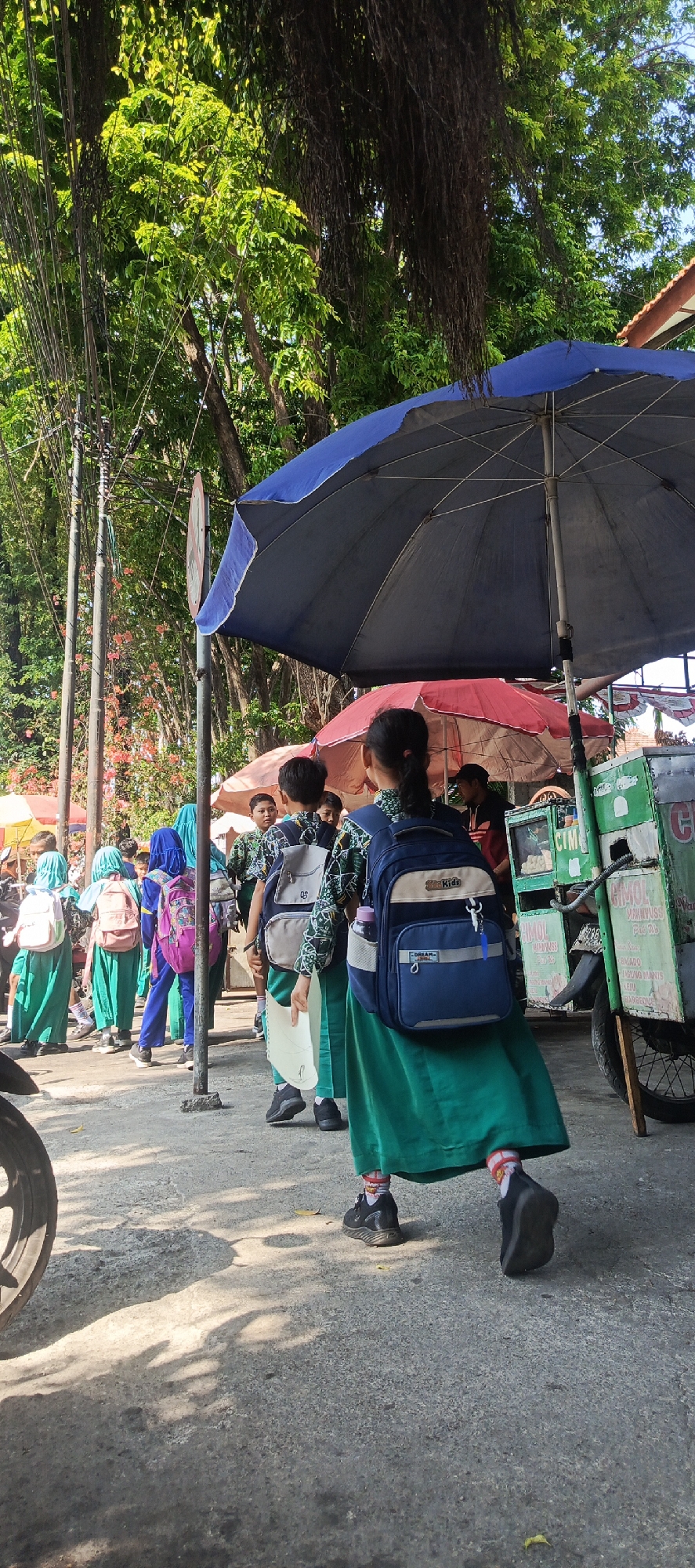 In Krian, Indonesia, a group of schoolchildren, mostly wearing green uniforms and carrying backpacks, are gathered on a concrete sidewalk. They appear to be waiting, possibly for transportation or at a market stall. The environment is outdoors, with lush green trees providing shade overhead and a street sign visible in the background. A street vendor's cart, covered by a large blue umbrella, is present, suggesting a commercial area. The light indicates it is daytime, with sunlight casting shadows on the ground. The overall scene depicts a typical day in a busy street in Indonesia, with children participating in daily activities.