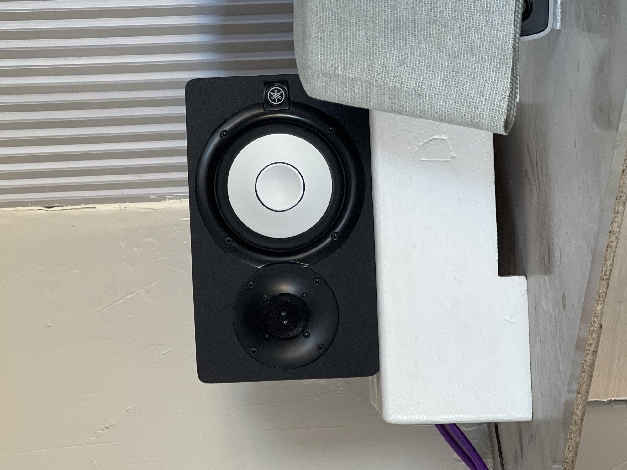 A black studio monitor speaker is positioned on a white styrofoam block. The speaker has two drivers: a smaller one on the left and a larger one on the right. The larger driver has a white cone and a black surround. The Yamaha logo is visible on the right side of the speaker. To the left of the speaker, purple cables are present. The speaker is situated on a countertop. Behind the speaker, a wall with vertical ribbed paneling is visible on the right, and a textured, off-white wall is on the left. A portion of a grey fabric object is visible to the right of the speaker. The image is taken indoors in a dimly lit environment. The location of Hazorasp, Uzbekistan is not depicted in the image.