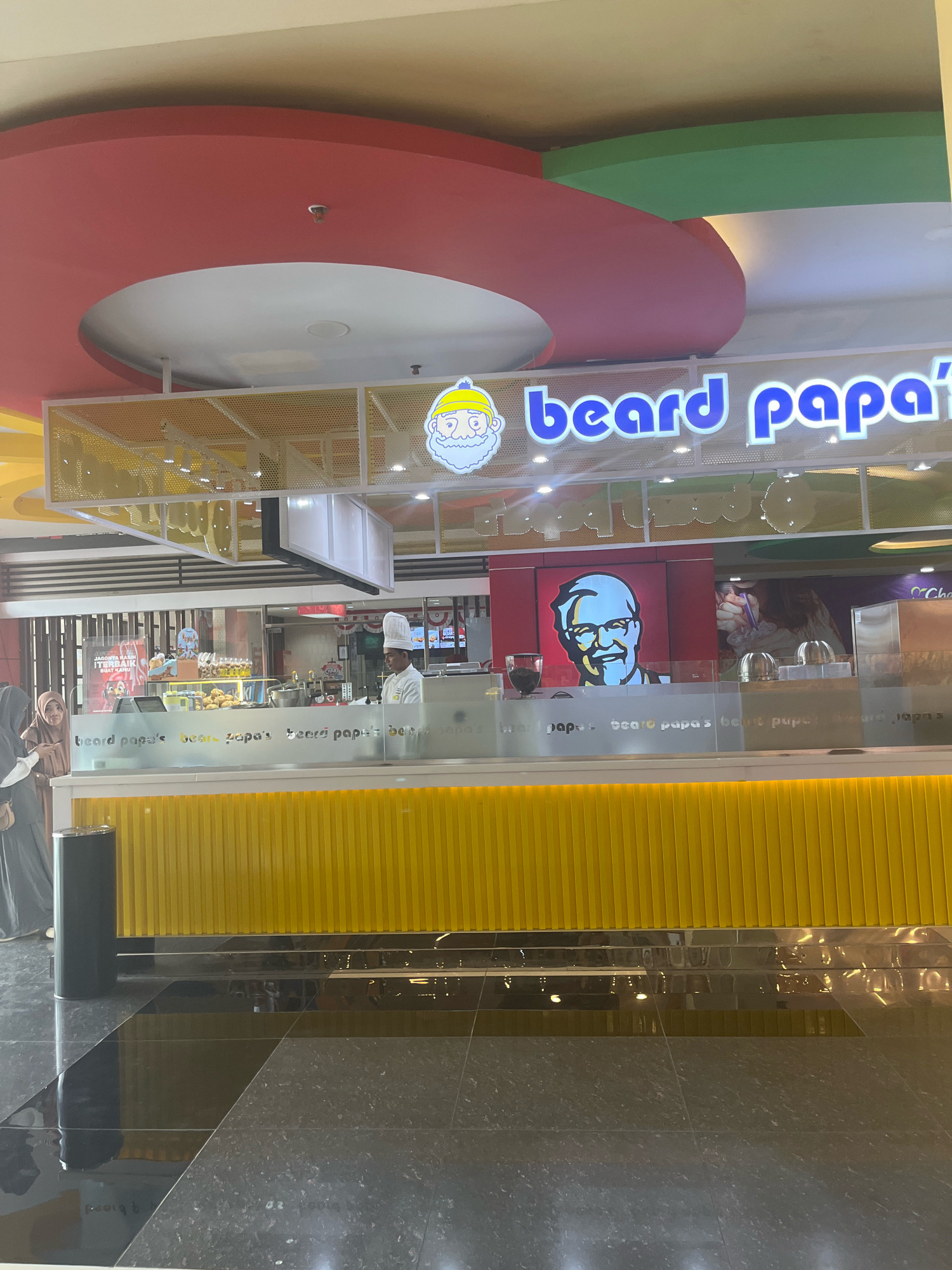 The image shows the exterior of a Beard Papa's store, a cream puff bakery, located in the city of Sengkang, Indonesia. The store features a bright yellow facade with the Beard Papa's logo prominently displayed above the counter. Inside the store, a chef in a white uniform is visible behind the counter, and customers can be seen browsing the selection of pastries. The store appears to be located within a shopping mall, as suggested by the polished floor and the ceiling design. The overall atmosphere is clean and inviting, with a focus on showcasing the products.