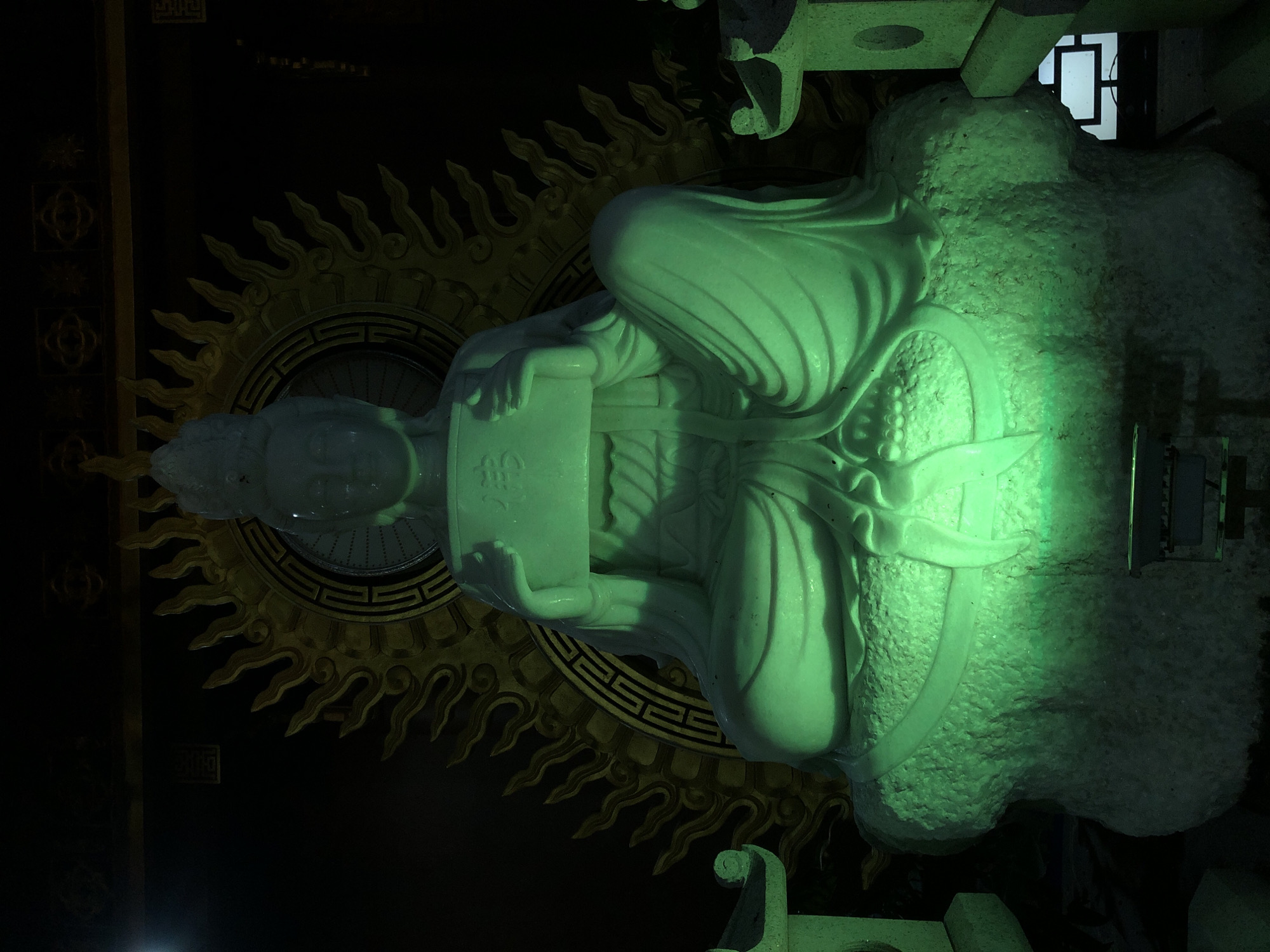 The image depicts a large, pale green statue of a seated figure, likely a deity or important religious figure, in what appears to be an indoor setting. The statue is illuminated by a bright green light, casting a strong glow on its surface. The figure is adorned with flowing robes, and it holds a rectangular object, possibly a book or tablet, in its hands. Behind the statue, a golden circular emblem with flame-like protrusions is visible, suggesting a religious or ceremonial context. The background is predominantly dark, with hints of architectural elements and what might be a window or screen in the upper right portion of the frame. The overall mood is solemn and reverent, intensified by the dramatic lighting. The location context indicates Mũi Né, Vietnam, which might suggest the statue is part of a temple or pagoda in that area. No specific time of day or weather conditions are discernible from the image. There is some visible text on the rectangular object held by the statue, though it is stylized and partially obscured by the lighting and the object's texture, making it difficult to decipher clearly.