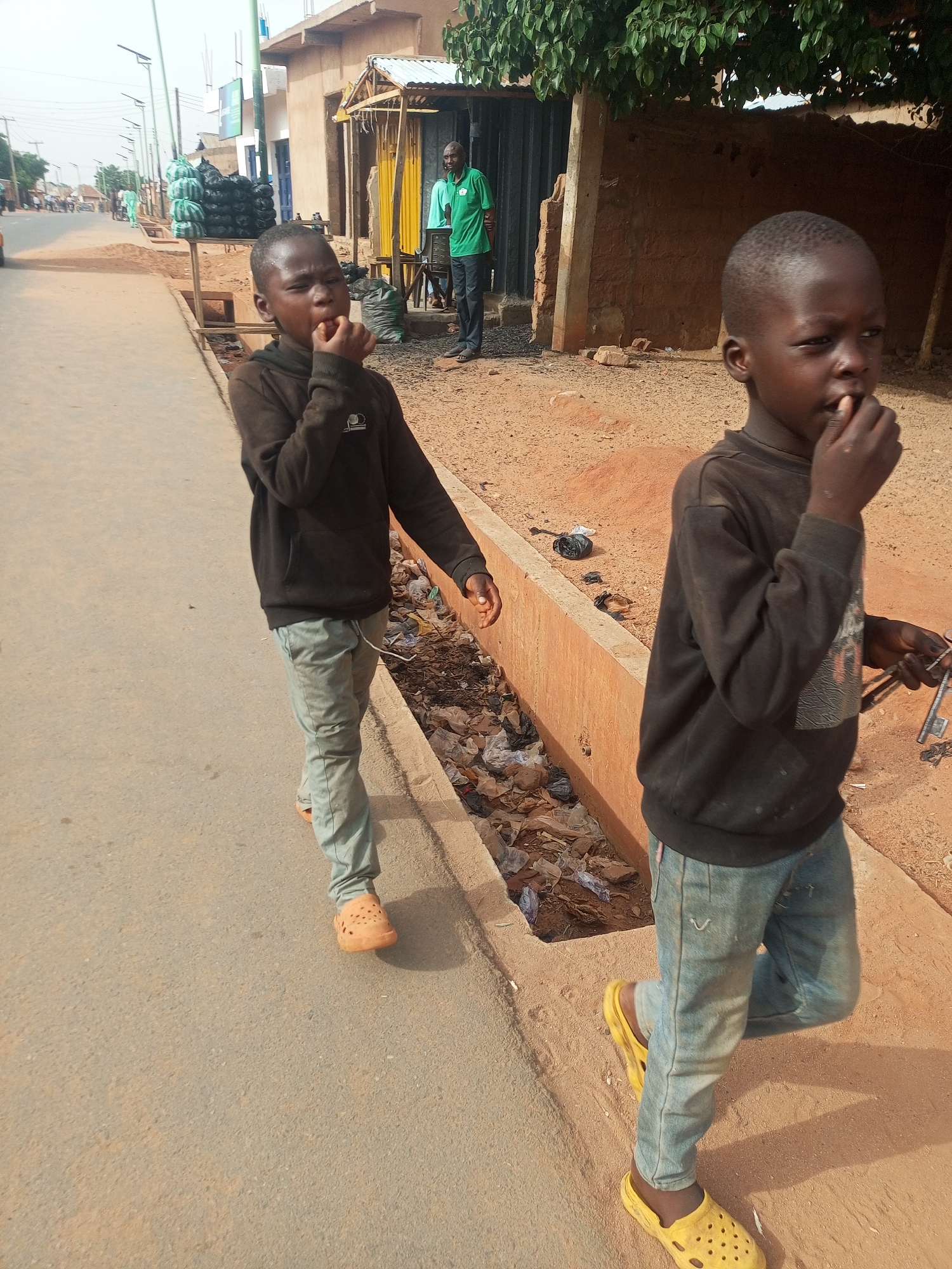 The image captures a street scene in Tempure, Nigeria, during the daytime. In the foreground, two young boys walk along a paved sidewalk. Both boys have dark skin and short hair, and their fingers are in or near their mouths, suggesting they might be eating or chewing on something.
The boy on the left is wearing a dark, long-sleeved hooded top, light grey pants, and orange Croc-style sandals. He is walking on the paved sidewalk, with a dirt patch and a litter-filled drainage ditch to his right. The boy on the right, slightly closer to the viewer and partially cut off by the frame, is wearing a similar dark, long-sleeved top, light blue jeans, and bright yellow Croc-style sandals. He holds a set of keys in his left hand.
In the mid-ground, behind the first boy, an adult male wearing a green polo shirt and dark trousers stands near a simple market stall or shop structure with a metal roof and yellow fabric. Black sacks, possibly filled with goods or refuse, are stacked on a wooden stand nearby. The background reveals a street stretching into the distance with more buildings and streetlights. The ground beside the sidewalk is predominantly unpaved earth, consistent with the specified location. The drainage ditch running parallel to the sidewalk contains visible plastic waste and other debris. The sunlight is bright, casting shadows, indicating it is likely midday or afternoon. No discernible text is visible on any signs or objects.