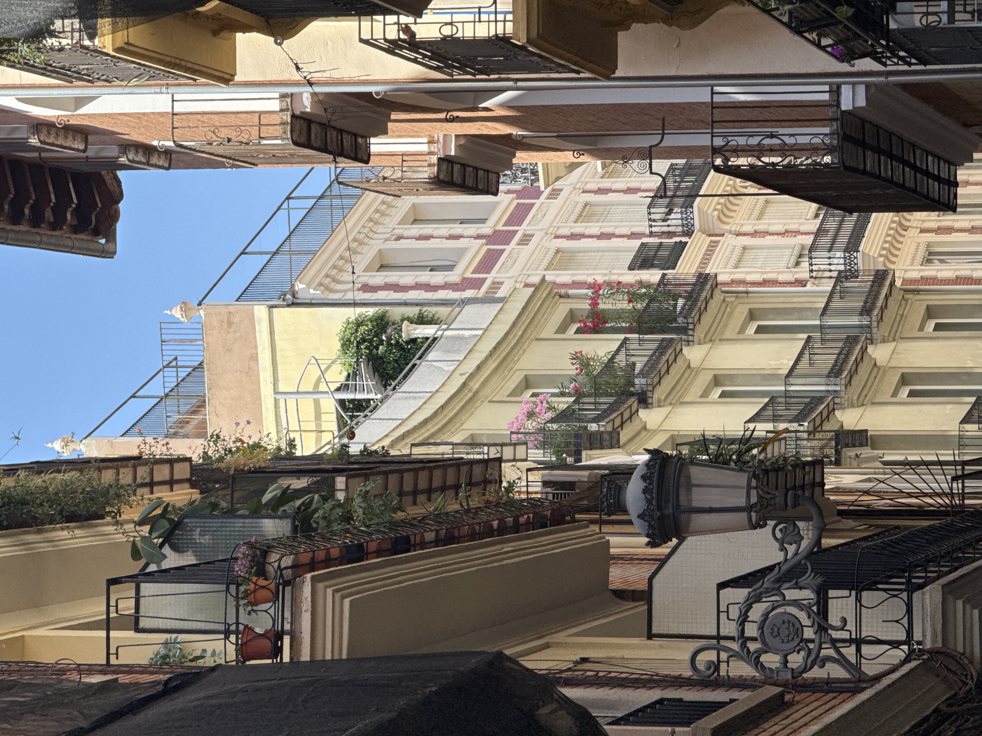 This image depicts a narrow street in Valencia, Spain, lined with ornate, multi-story buildings. The architecture features balconies with wrought-iron railings, often adorned with potted plants and flowers, such as pink blossoms visible on one of the balconies. Many of the buildings are painted in warm tones of yellow and ochre, with contrasting architectural details in brick and white stucco. A vintage-style lamppost with intricate metalwork is prominently featured in the foreground, extending from a building's facade. Above, spiral staircases and rooftop terraces are visible, hinting at the layered urban landscape. The sky is clear and blue, indicating a sunny day. There are no people visible in the image, and the scene is static and architectural. The overall impression is one of a charming, historic European street.