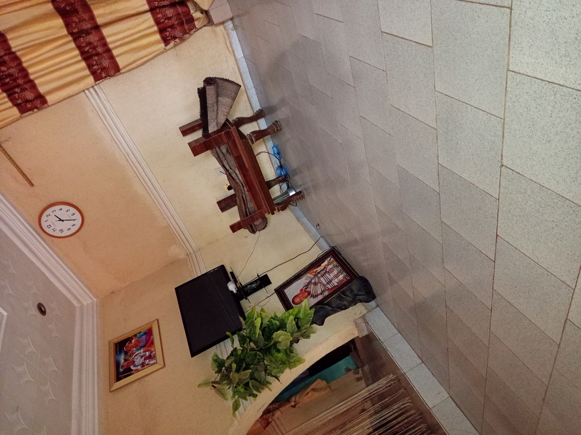 A tilted medium shot captures an indoor scene with a light-colored tiled floor. In the upper left, a framed photograph of two individuals hangs on a light-colored wall with a decorative ceiling. To the right of the photograph, a round clock with a white face and black numbers is mounted on the wall. A dark flat-screen television is mounted on the wall below the clock. To the left of the television, a large artificial plant with green leaves is placed on a dark, curved stand. In the lower left, a framed photograph of an individual in traditional attire leans against a dark, abstract sculpture. In the center of the image, a wooden bench is overturned on its side, revealing rolled-up fabric on its underside. A blue plastic water bottle is visible on the floor beneath the overturned bench. To the right of the bench, a gold and burgundy striped curtain hangs by a window. The overall scene depicts an interior space with decorative elements and furniture.