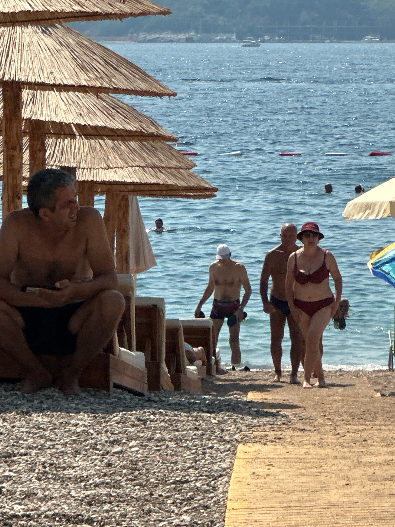 The image captures a sunny day on a beach, likely in Budva, Montenegro, with the Adriatic Sea serving as the backdrop. Several people are visible on the shore and in the water. In the foreground, a man with graying hair is seated under a thatched umbrella, looking to his left. Further down the beach, three adults are walking along the water's edge; a woman in a red bikini is in the lead, followed by two men. One man wears dark swim trunks, and the other wears patterned swim shorts and a white cap. In the water, several other individuals can be seen swimming or wading, with buoys marking a designated area. The beach itself is a mix of pebbles and sand, with wooden pathways laid out. The sea is a vibrant blue, reflecting the bright sunlight. The scene evokes a typical summer vacation atmosphere.