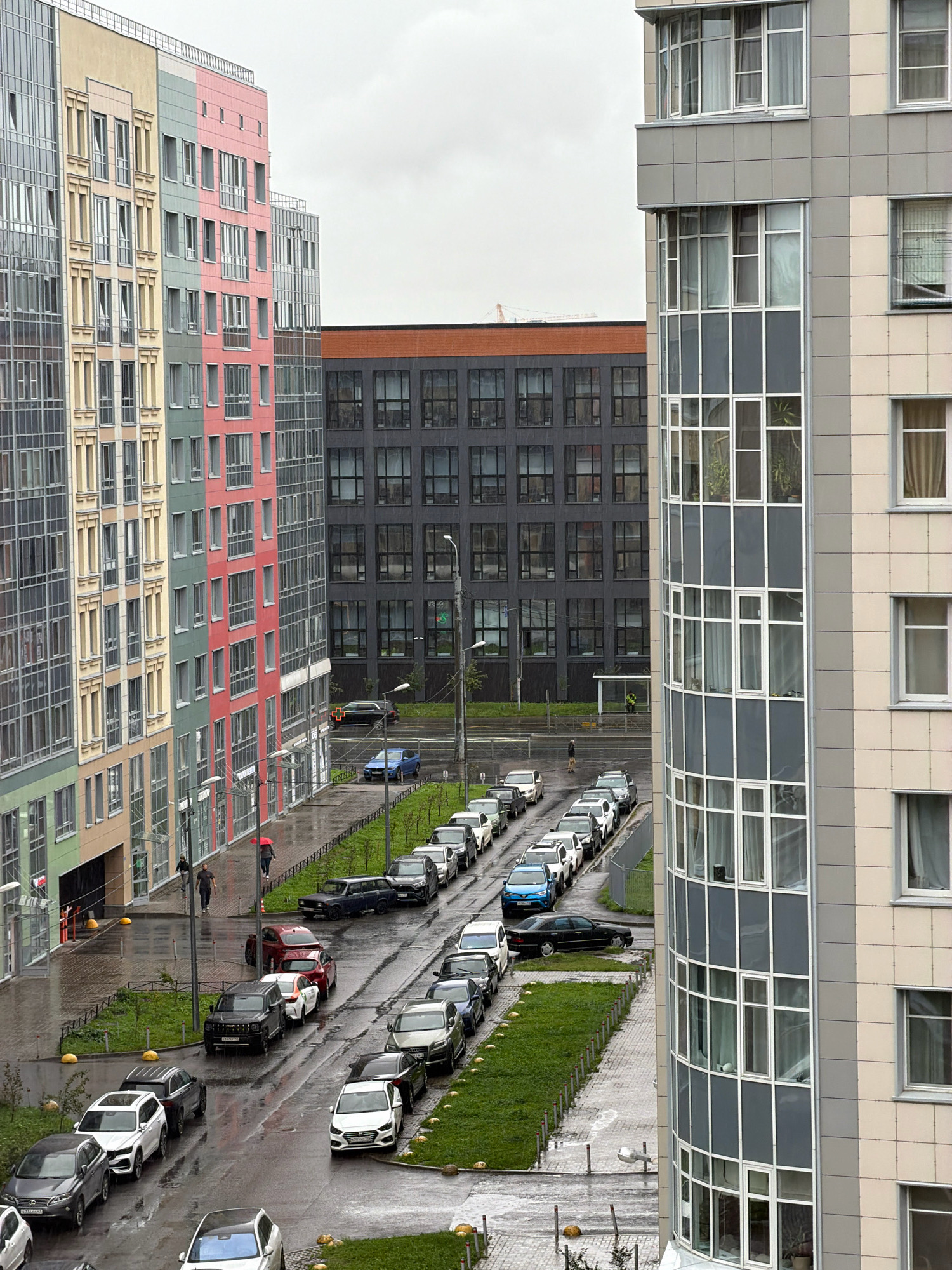 The image depicts an urban scene with modern residential buildings and parked cars on a wet street. The buildings feature a combination of colorful and grey facades with numerous windows. A street with parked cars runs between the buildings. Several people are visible walking on the sidewalk, with one person holding a red umbrella. The ground is wet, indicating recent rain. In the background, a darker, larger building with many windows is visible. The sky is overcast.