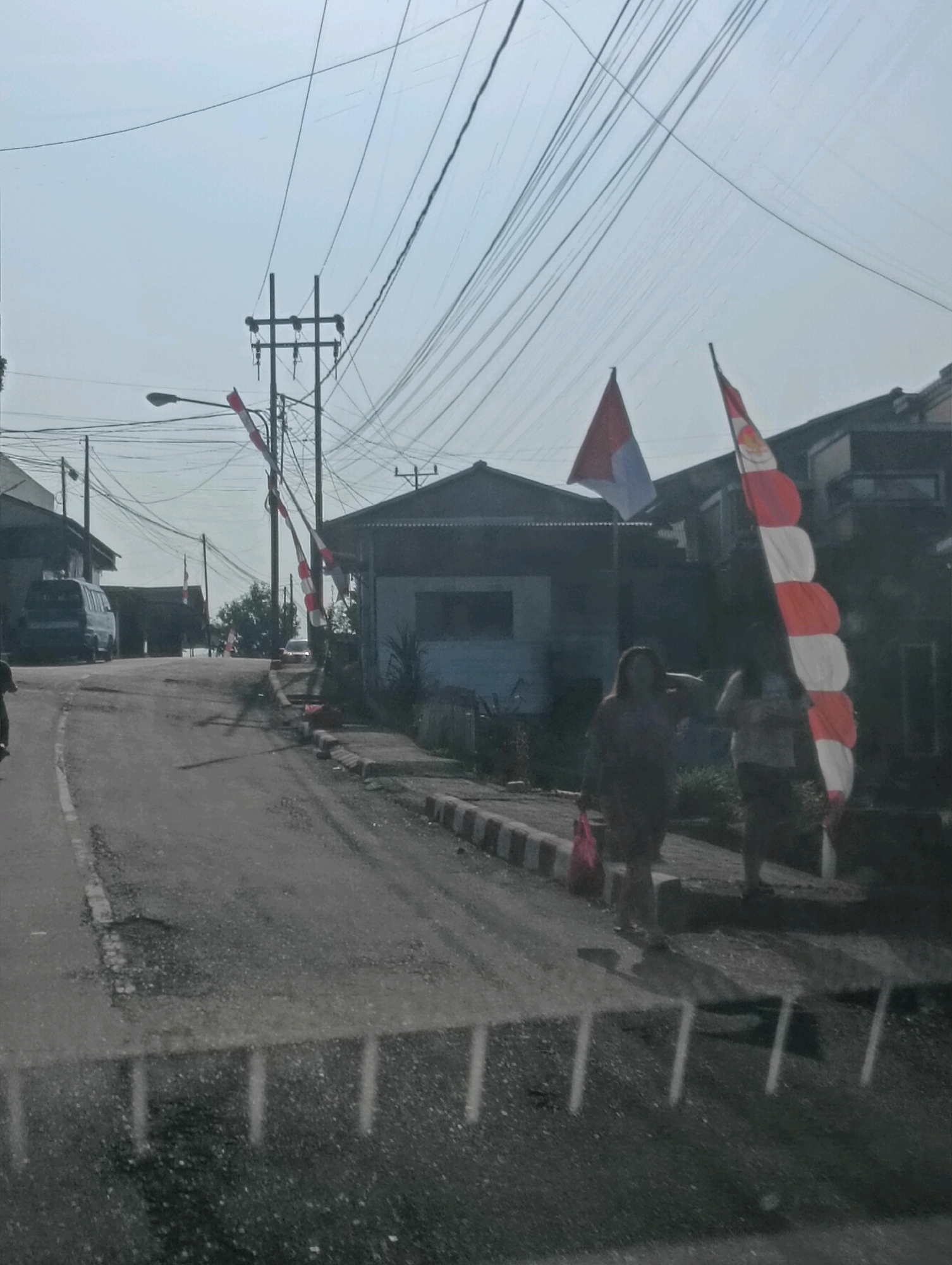 The image depicts a street scene in Tomohon, Indonesia, with a slightly hazy sky and numerous overhead power lines dominating the upper portion of the frame. A paved road, showing signs of wear and tear with visible potholes, curves through the scene. On the right side of the road, a raised sidewalk with red and white striped barriers is present, with two women walking along it. One woman, wearing a dark patterned shirt and carrying a pink bag, is on the left. The other woman, wearing a lighter colored shirt, is to her right. Behind them, a red and white striped flag, likely representing the Indonesian flag, is prominently displayed on a pole. Houses with dark roofs are visible along the sides of the street. A blue van is parked on the left side of the road further up the street. The overall impression is of a moderately busy residential or commercial street in Indonesia, likely during daytime.