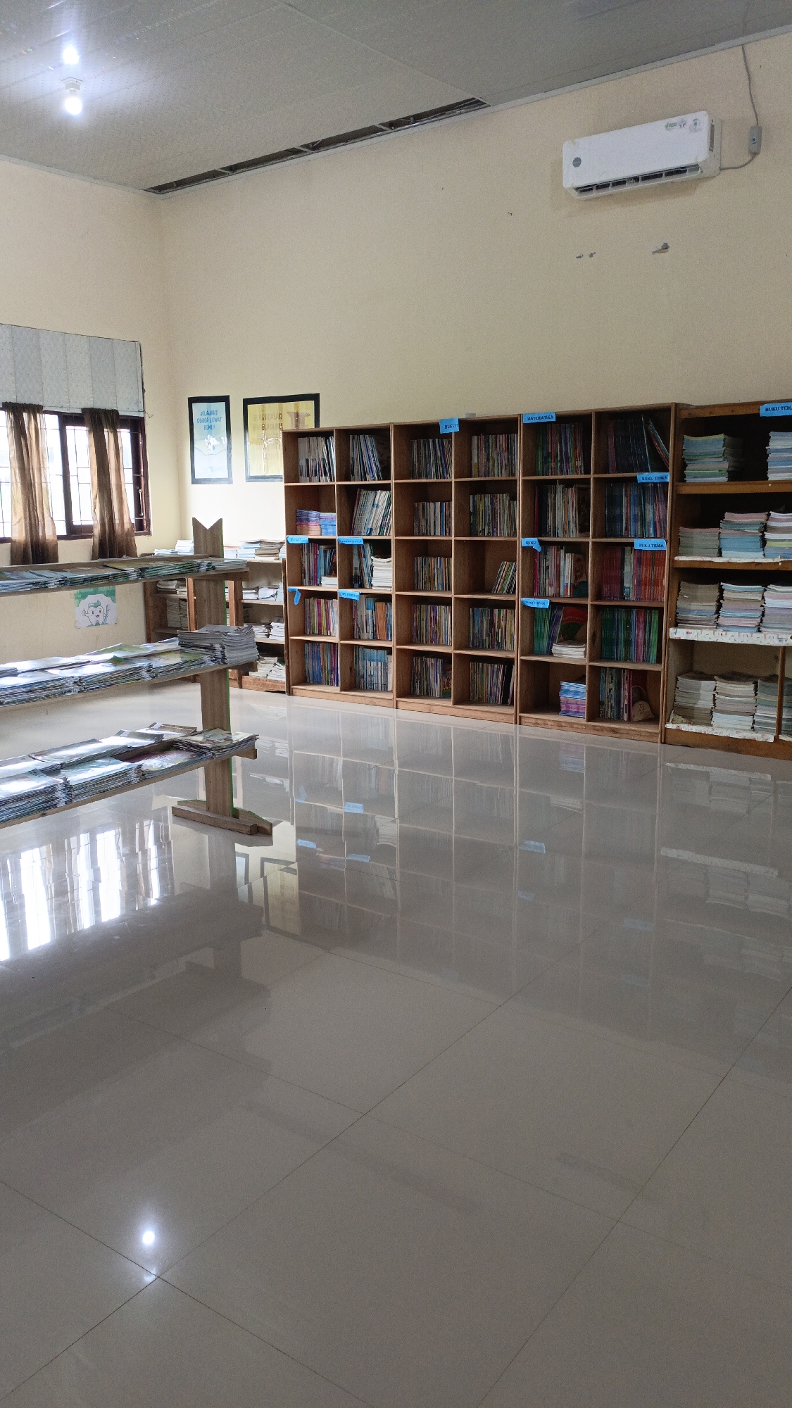 The image depicts the interior of a library, likely in Saring Sungai Binjai, Indonesia. The room is illuminated by overhead lights and natural light filtering through windows covered by brown curtains. The walls are painted a pale yellow, and the ceiling appears to be acoustic tile.
The primary features of the library are multiple wooden bookshelves filled with books. One large set of bookshelves is positioned against the back wall, featuring numerous compartments stocked with books of various sizes and colors. Some shelves have blue labels, indicating categorization. To the right, a narrower set of bookshelves also contains books. In the foreground, there are two long, low wooden shelving units displaying stacks of books that appear to be laid flat. The floor is tiled with large, light-colored tiles that reflect the light and the objects in the room.
On the wall above the bookshelves, an air conditioning unit is mounted. To the left, there are two framed pictures hanging on the wall. One appears to be a poster with text and an illustration, and the other is a framed picture with some text.
The scene is quiet and orderly, suggesting a functional library space. There are no people visible in the image. The overall impression is that of a well-maintained educational or public facility.