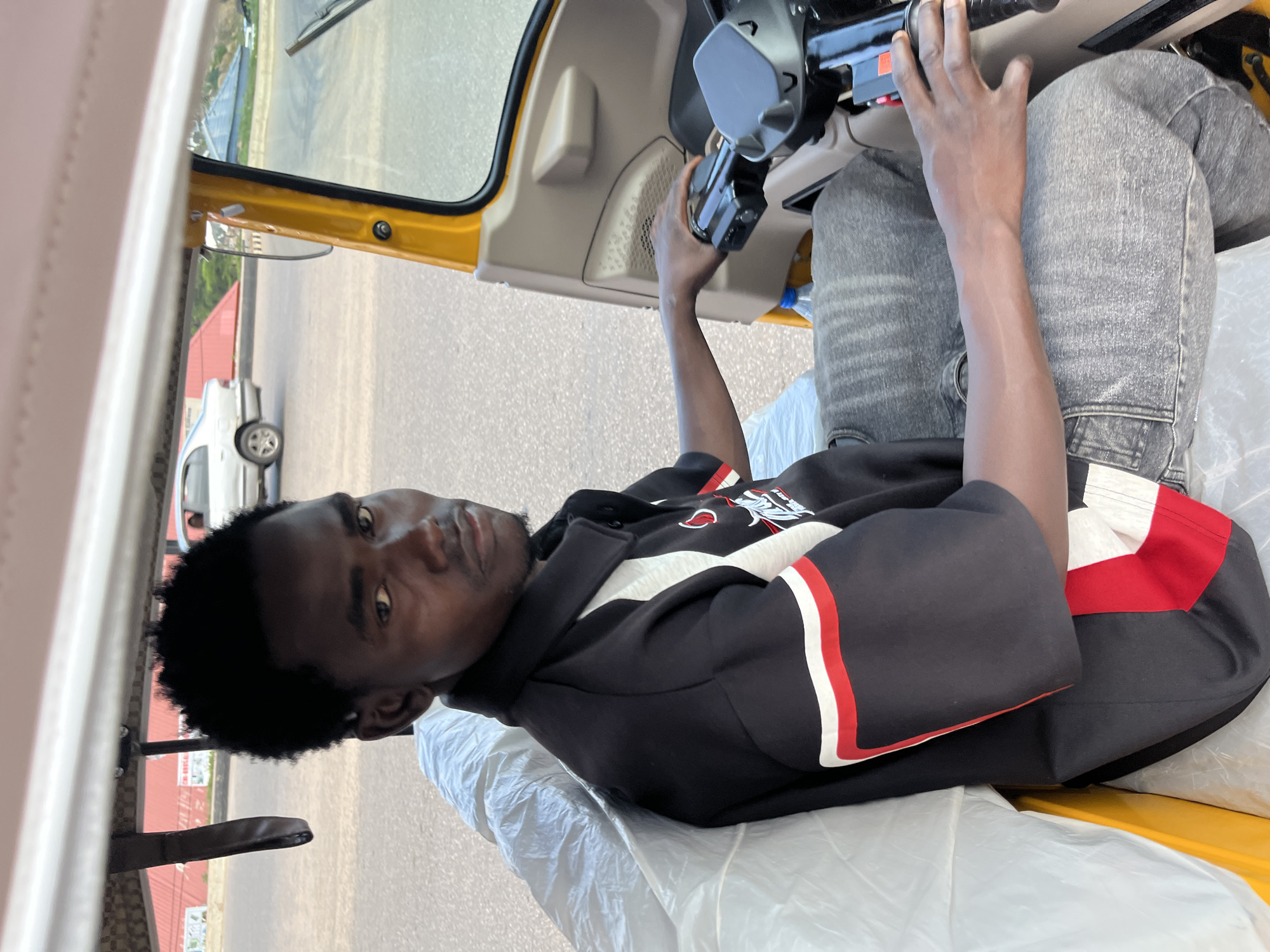 A man is seated in the driver's seat of a yellow three-wheeled vehicle, commonly known as a "keke." He is wearing a black long-sleeved shirt with red and white stripes on the sleeves and chest, and grey denim jeans. The man has dark skin and short, curly black hair. He is looking towards the camera with a neutral expression. His right hand is resting on the steering column. The interior of the vehicle appears to be simple, with beige plastic panels. A white plastic sheet is covering the seat next to him. Outside the vehicle, a paved road and some buildings can be seen. The lighting suggests it is daytime. The scene is set in Gura Top, Nigeria.